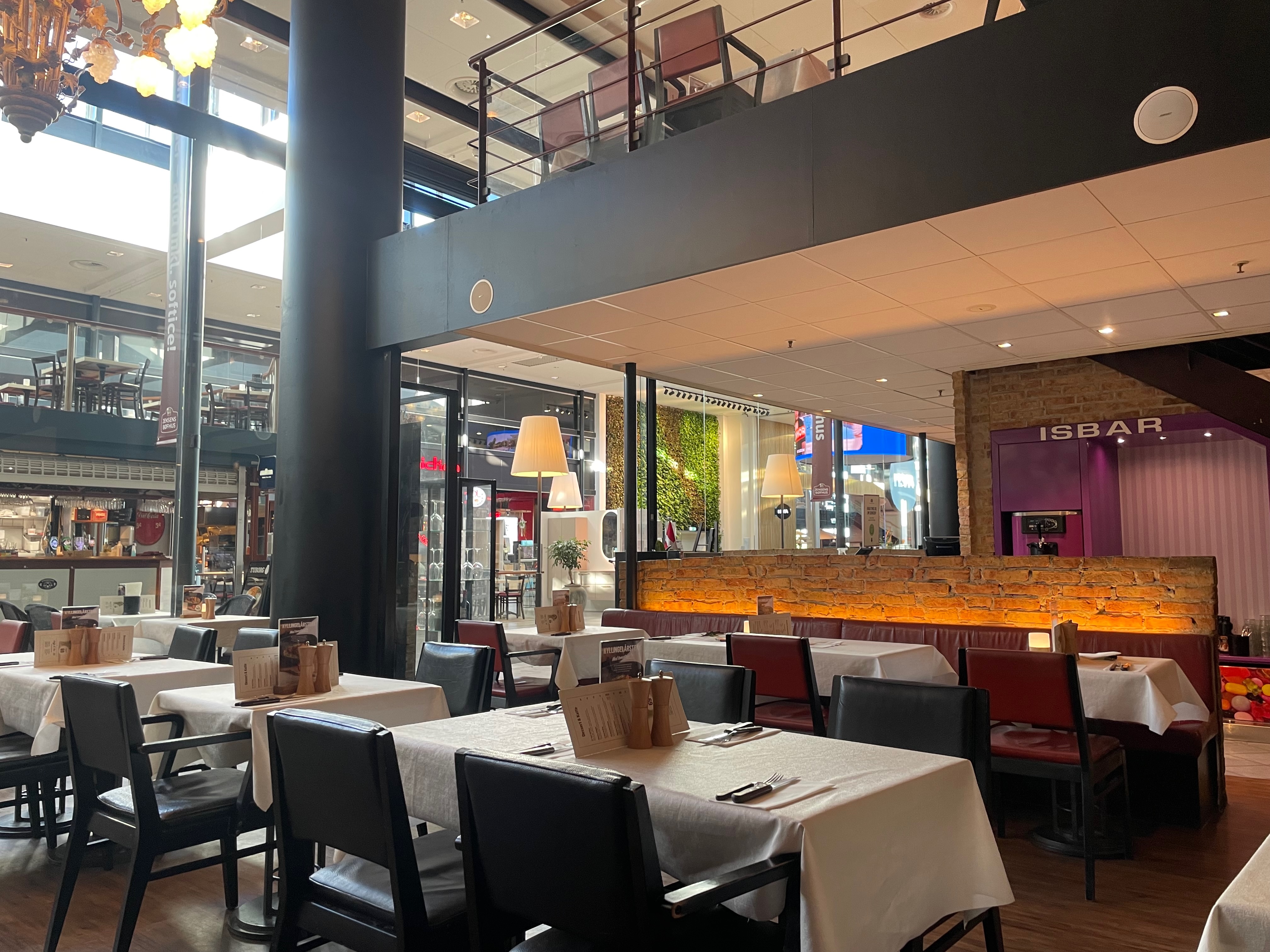 Jensens Bøfhus Kolding review restaurant interior inside Kolding Storcenter, showing white tablecloth tables, black and red chairs, warm lighting, brick wall seating, and the nearby ISBAR area