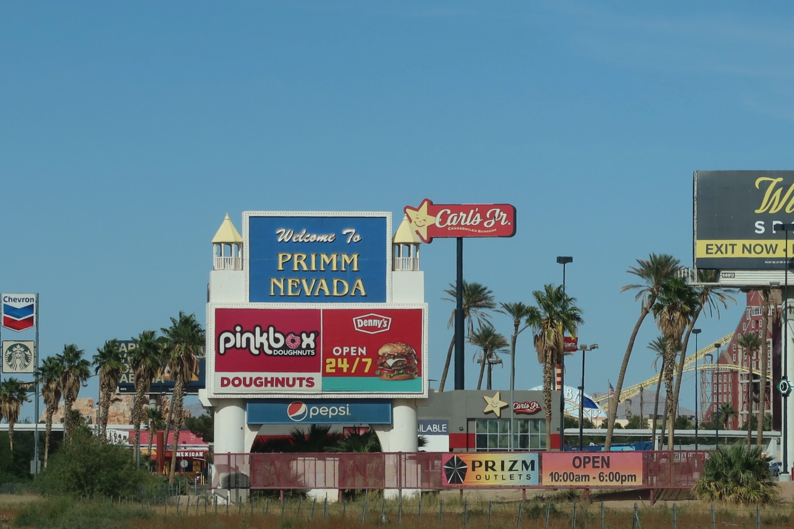 Welcome to Primm Nevada roadside sign featuring Pinkbox Doughnuts, Denny’s open 24/7, Pepsi branding, and Prizm Outlets, surrounded by palm trees under a clear desert sky.