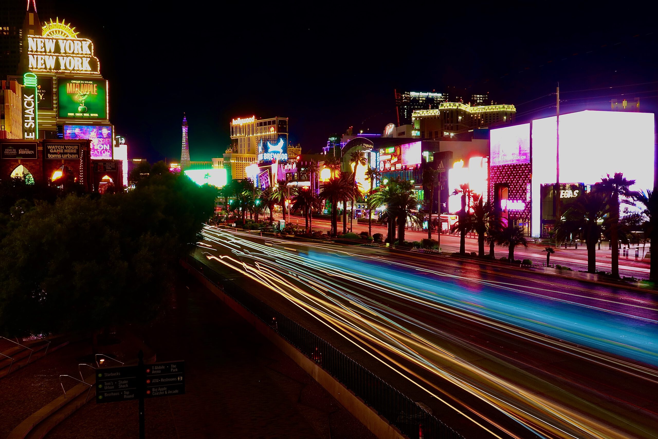 Night view of the Las Vegas Strip with long-exposure light trails from passing cars, palm-lined boulevard, bright neon billboards, and iconic hotels including New York-New York glowing against the dark desert sky.
