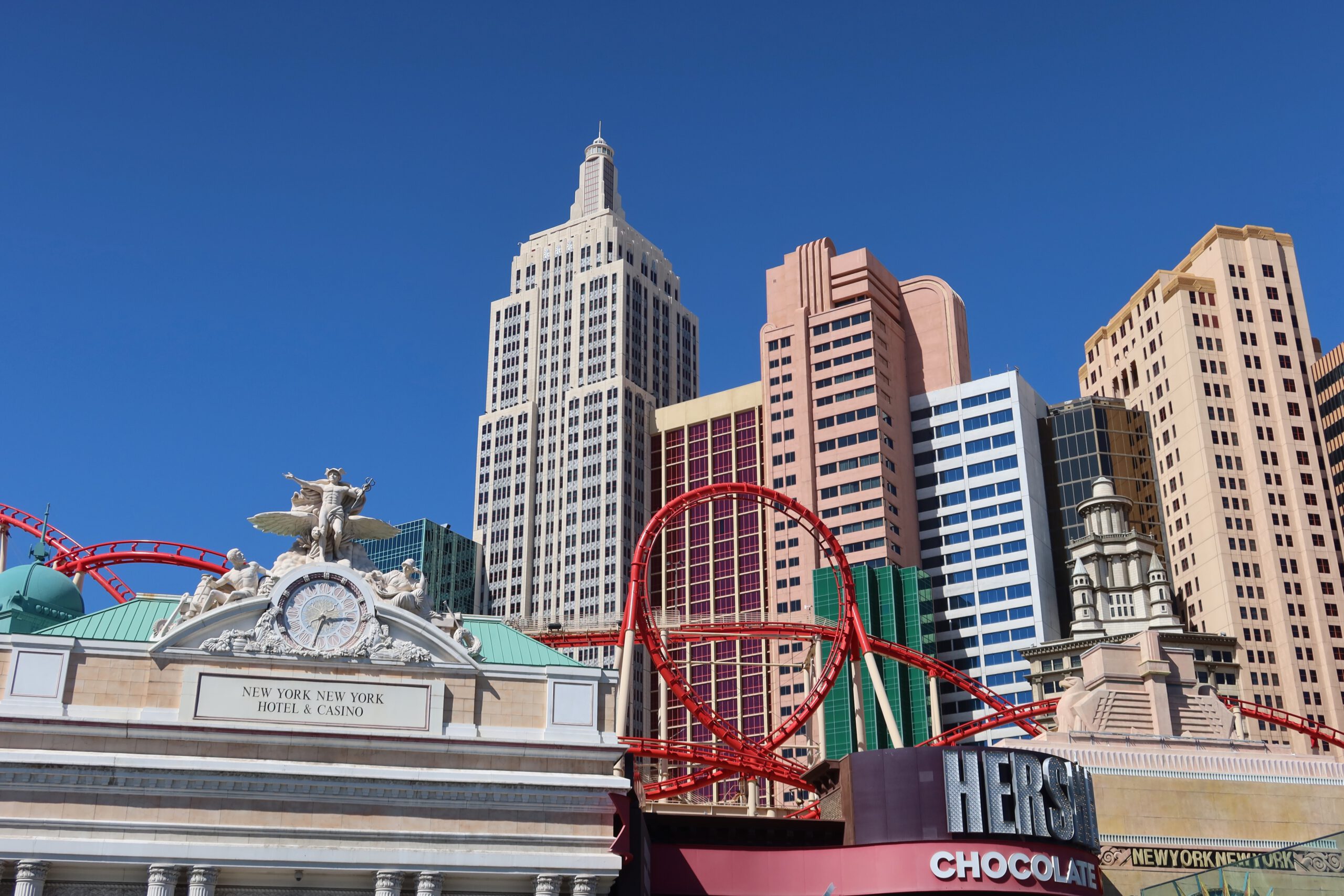 The Big Apple Coaster at New York-New York Hotel and Casino in Las Vegas, with the red roller coaster loop rising in front of a New York–inspired skyline under a clear blue sky.