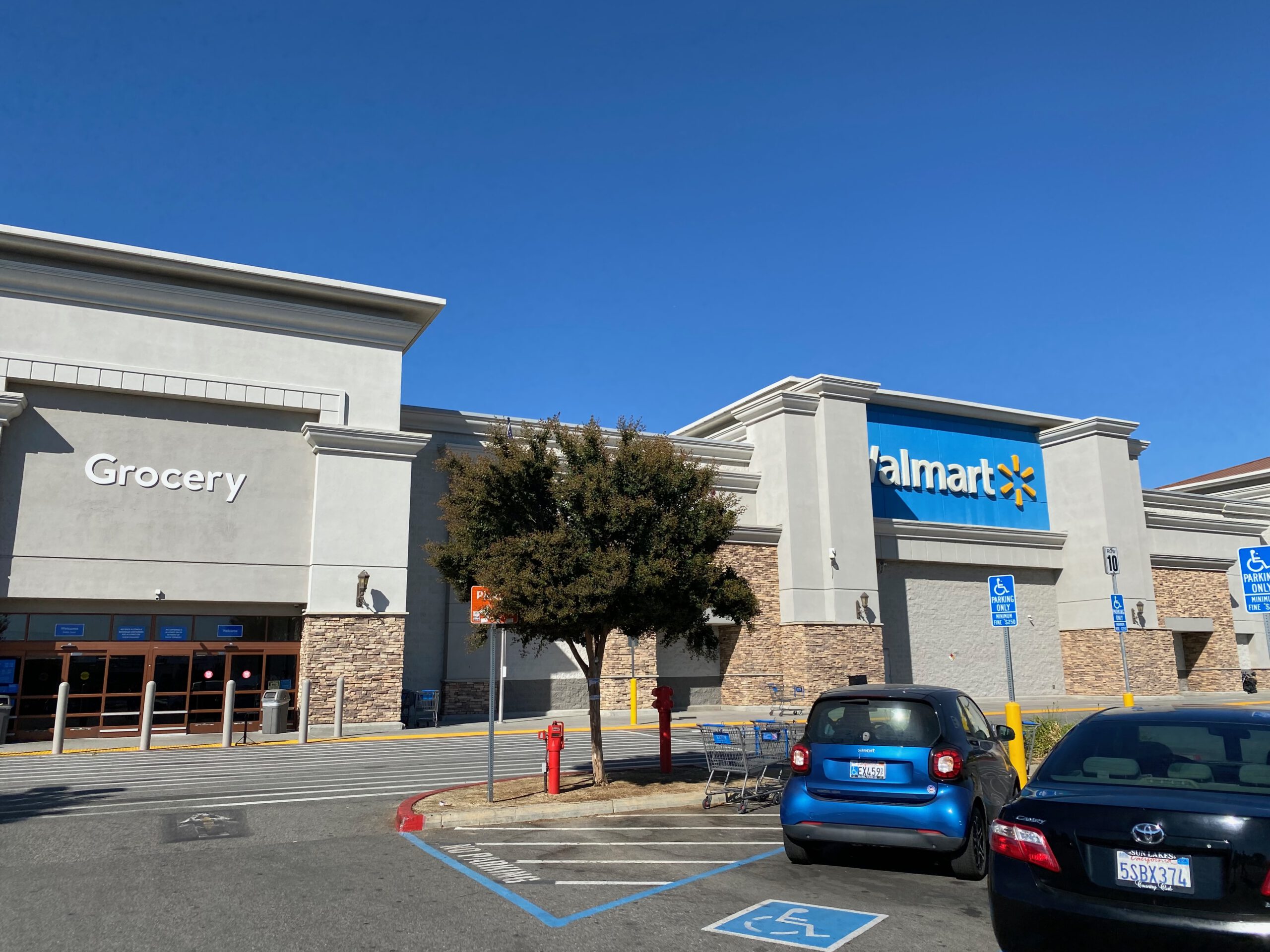Front exterior of Walmart Supercenter Beaumont California with large Walmart logo, grocery entrance signage, wide parking lot, blue California sky, and shopping carts visible near the entrance.