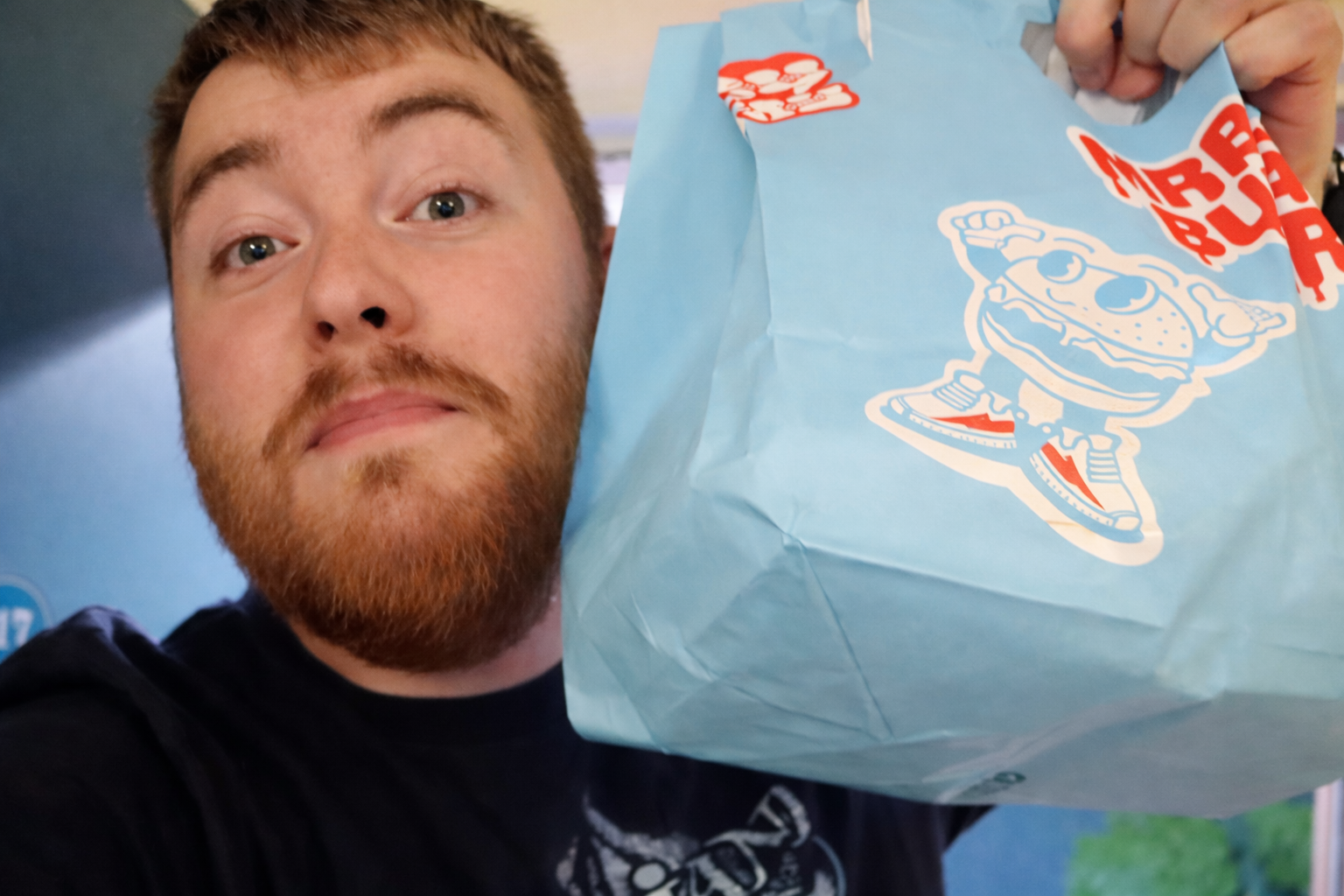 Timo van den Berg aka Timeuw holding a MrBeast Burger takeout bag in San Francisco, showing the light blue MrBeast Burger branding and mascot during a food delivery experience.