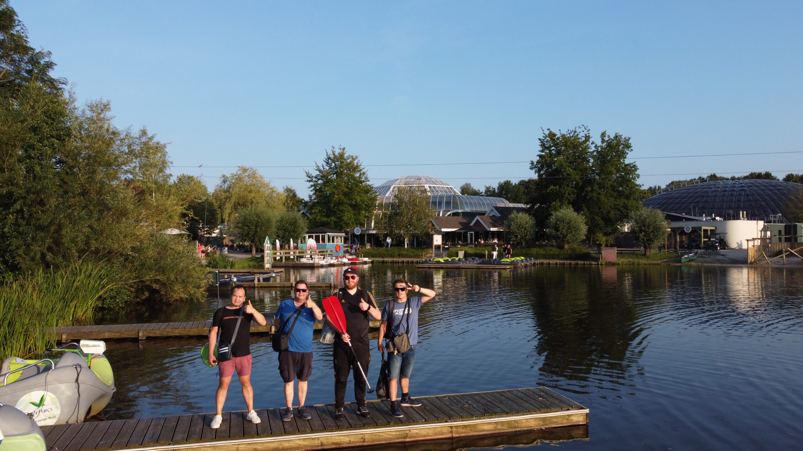 Center Parcs De Huttenheugte Day Visit at the lake in Dalen Drenthe, Chris and Rogier from AMFVLOGS together with Peter Smit and Timeuw Timo van den Berg standing on a wooden dock with paddle boats and the Aqua Mundo dome visible in the background during golden hour.