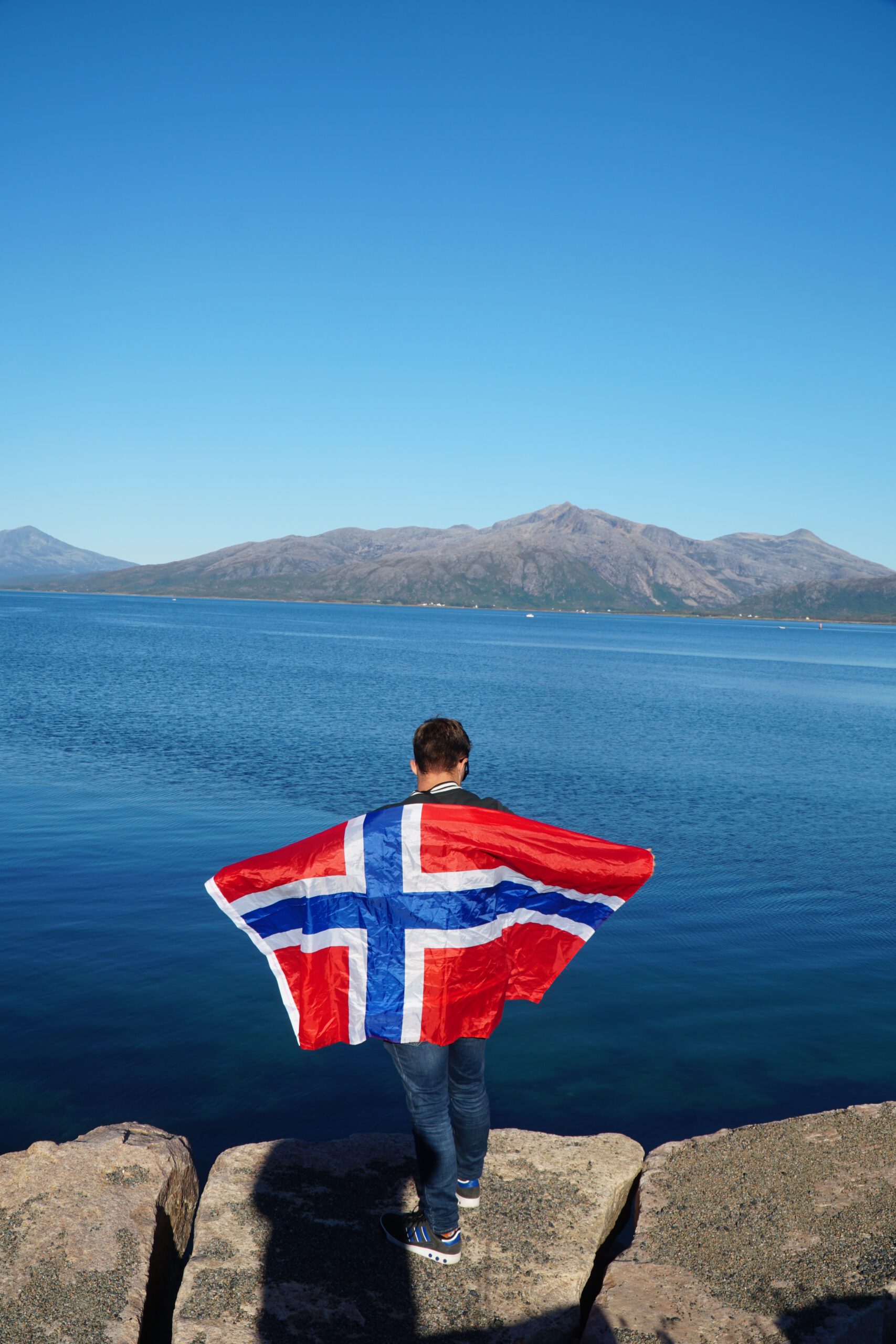 Peter Smit holding the Norwegian flag near Lødingen Brygge Hotel Norway, overlooking the fjord with calm blue water and mountain landscape in Northern Norway