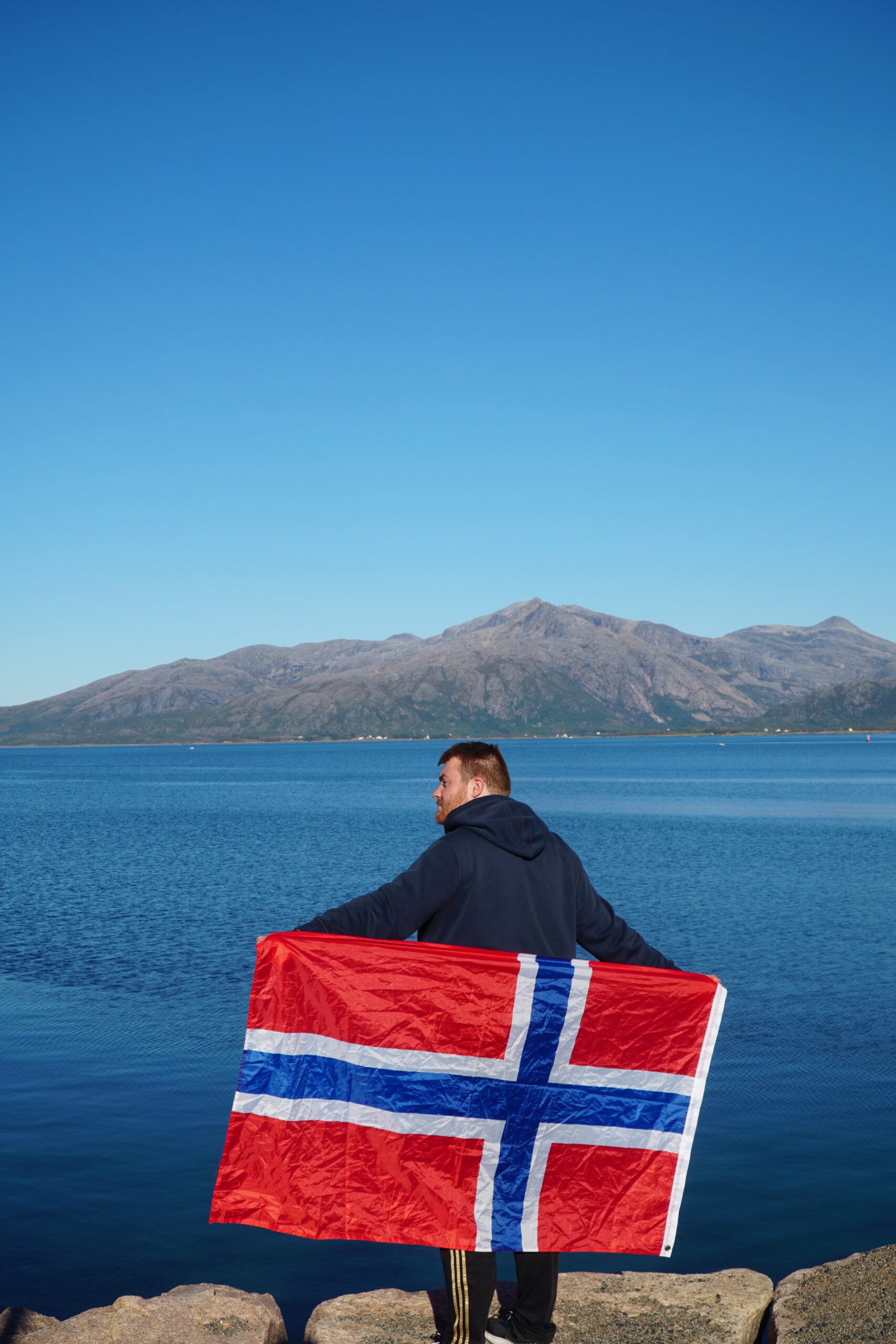 Timo van den Berg Timeuw holding the Norwegian flag near Lødingen Brygge Hotel Norway, standing by the fjord with mountains and clear blue water in Northern Norway