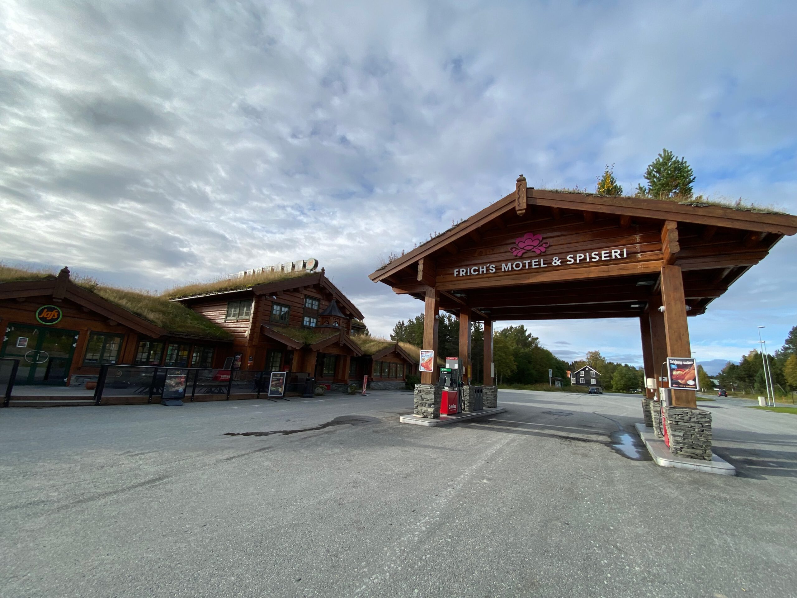 Oppdalsporten Fagerhaug entrance along the E6 highway in Norway, featuring Frich’s Motel & Spiseri wooden log buildings with grass roof architecture and a Circle K Automat fuel pump under a large timber canopy, surrounded by Norwegian mountain landscape and cloudy sky.