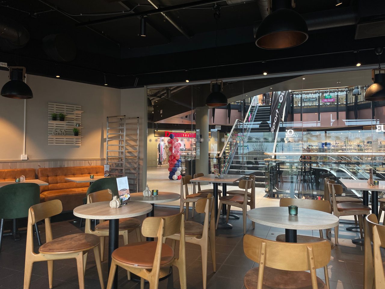 Cafe Sjøsiden in Mosjøen, showing the cozy indoor seating area with tables, chairs, booth seats, and a view toward the Alti Sjøsiden shopping center.