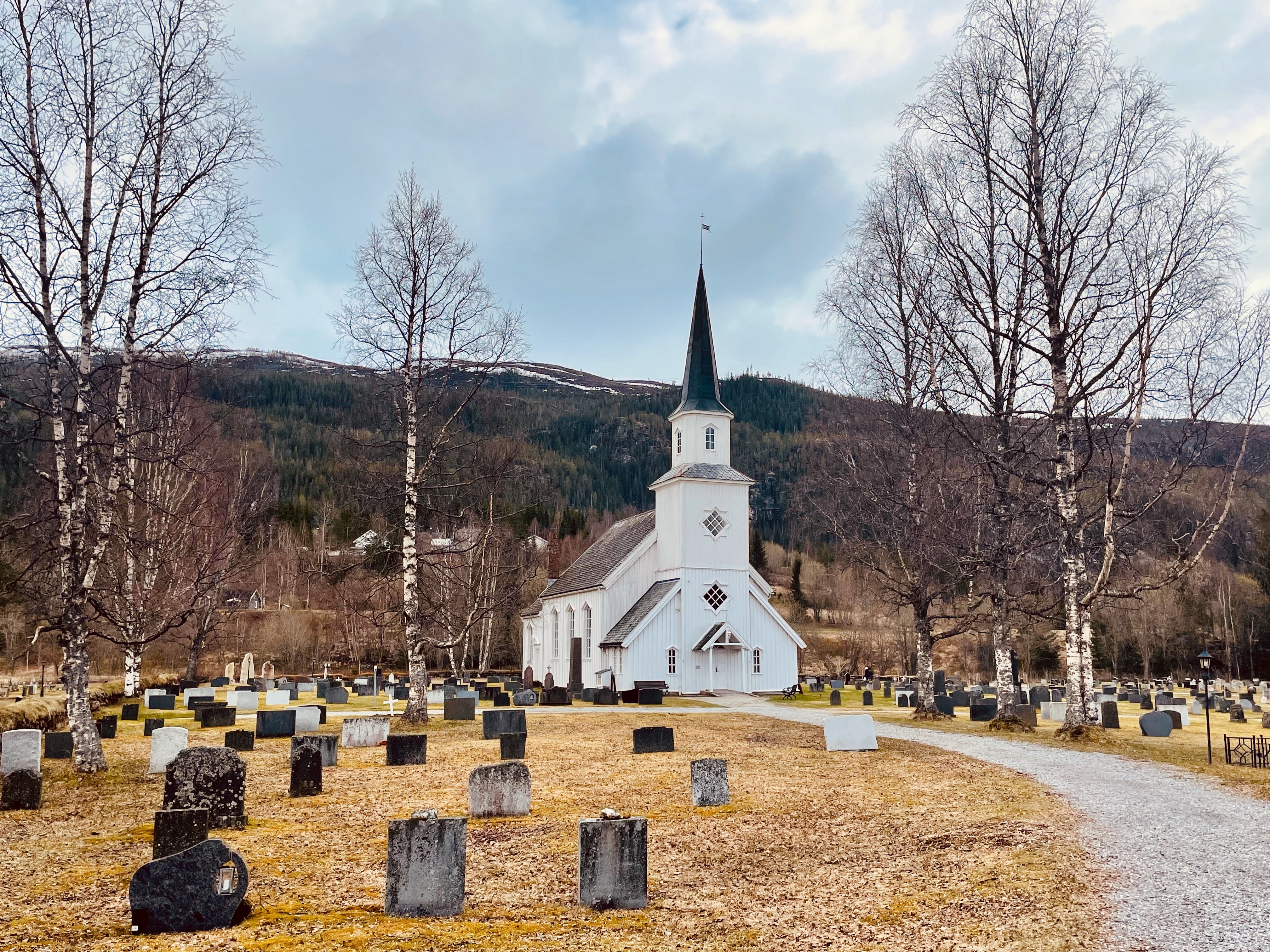 Grane Church in Grane near Trofors, a white wooden church surrounded by graves, birch trees, a gravel path, and mountain scenery in Northern Norway.