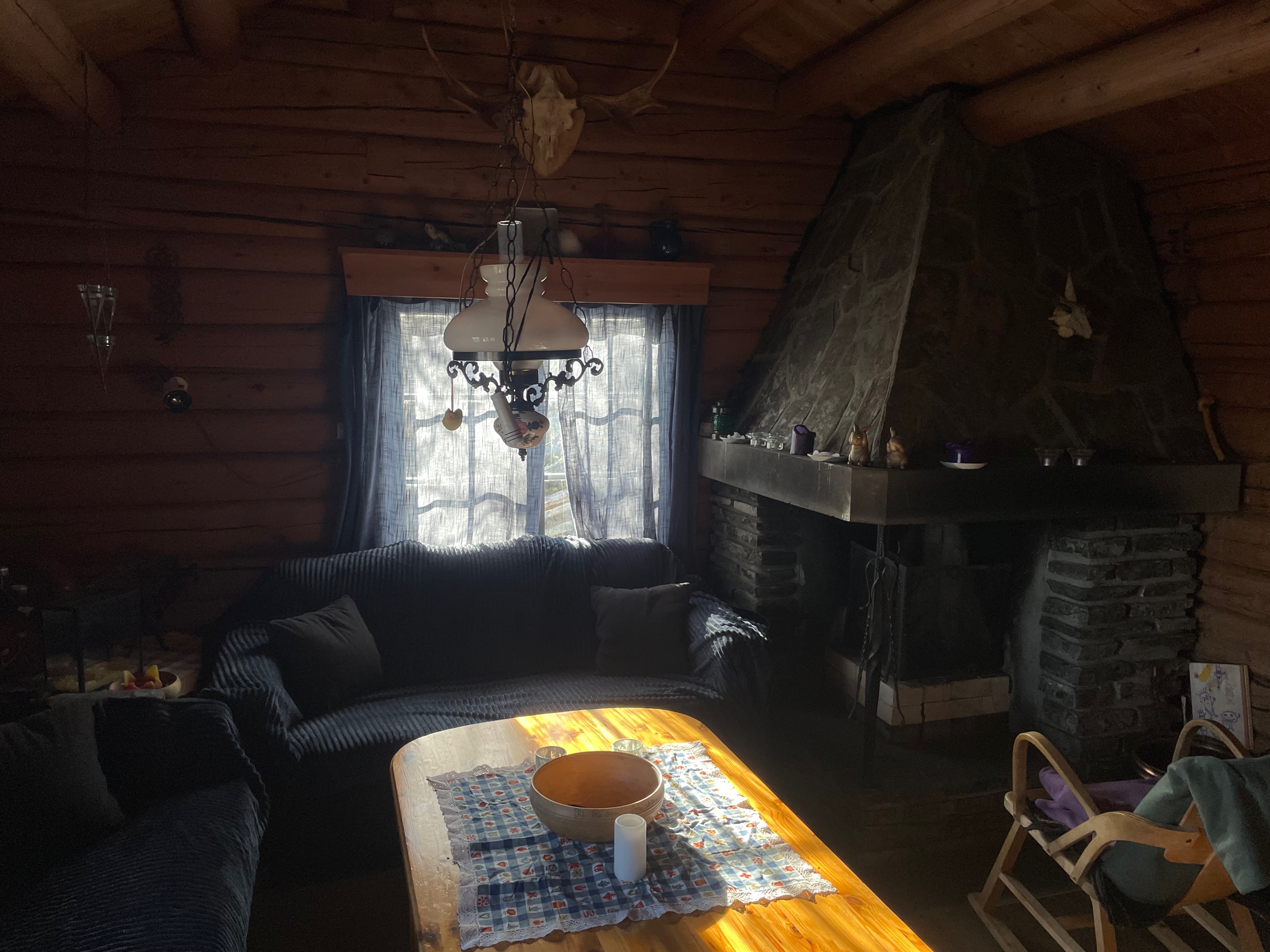 Per’s stuggu Sør-Fron rustic living room with dark wooden walls, blue sofas, stone fireplace, wooden coffee table, antler decor, hanging lamp and cozy old cabin interior.
