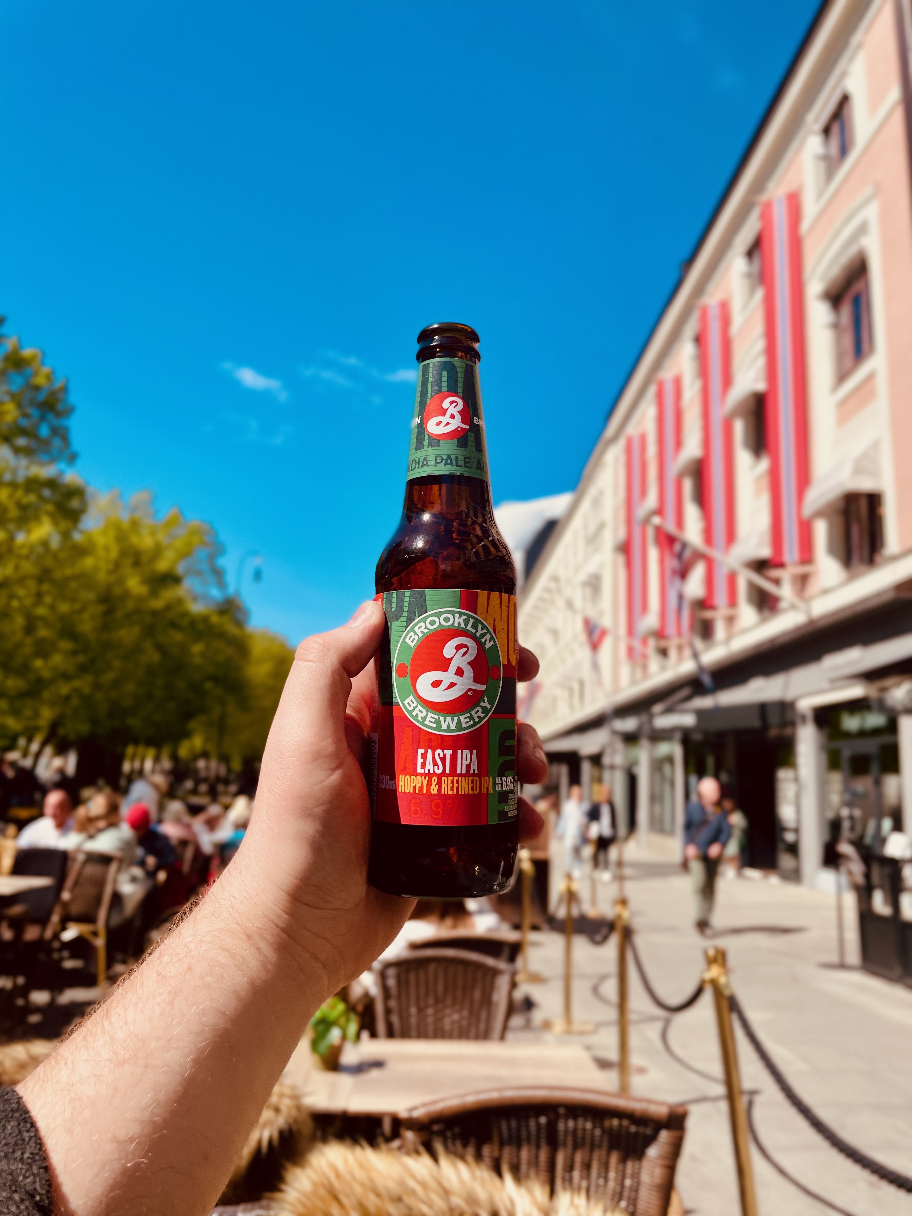 Brooklyn Brewery East IPA bottle held in front of Egon Karl Johan terrace on Karl Johans gate in Oslo, with sunny outdoor seating and city buildings in the background.