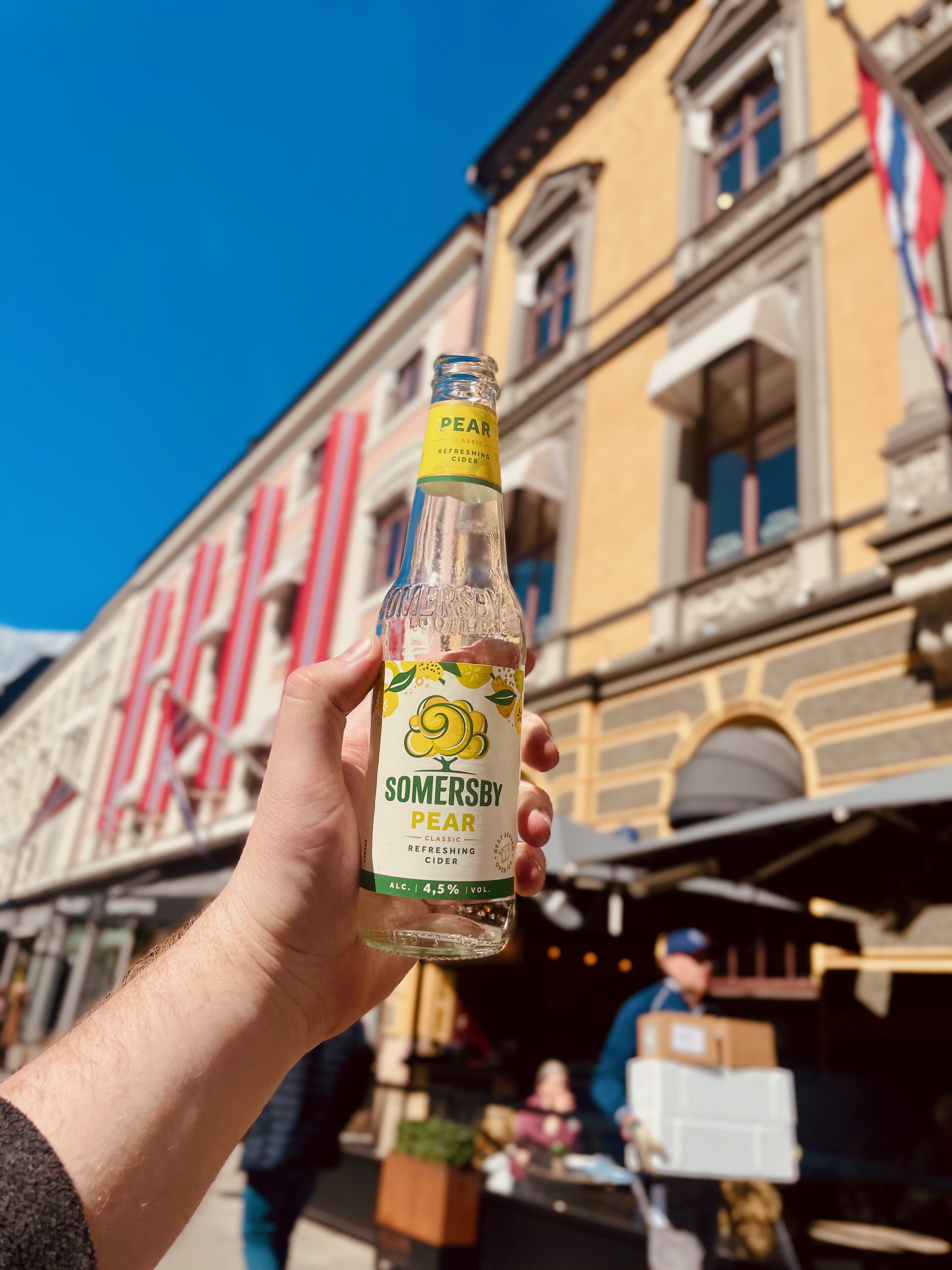 Somersby Pear cider bottle held in front of Egon Karl Johan restaurant in Oslo, with the yellow facade, terrace seating, umbrellas, and blue sky in the background.