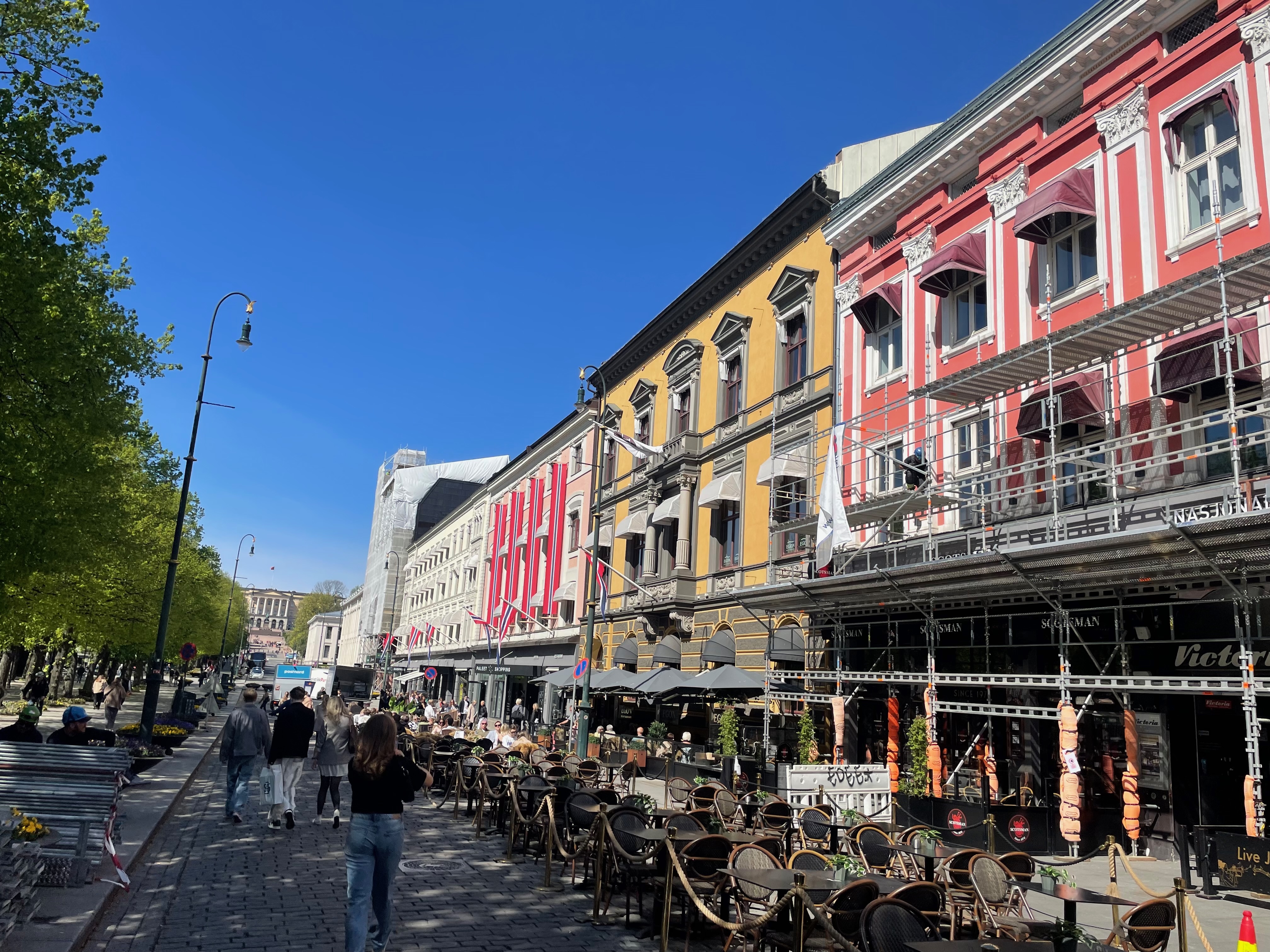 Egon Karl Johan on Karl Johans gate in Oslo, showing the sunny restaurant terrace, historic colorful facades, outdoor seating, Norwegian flags, and people walking through the city center.