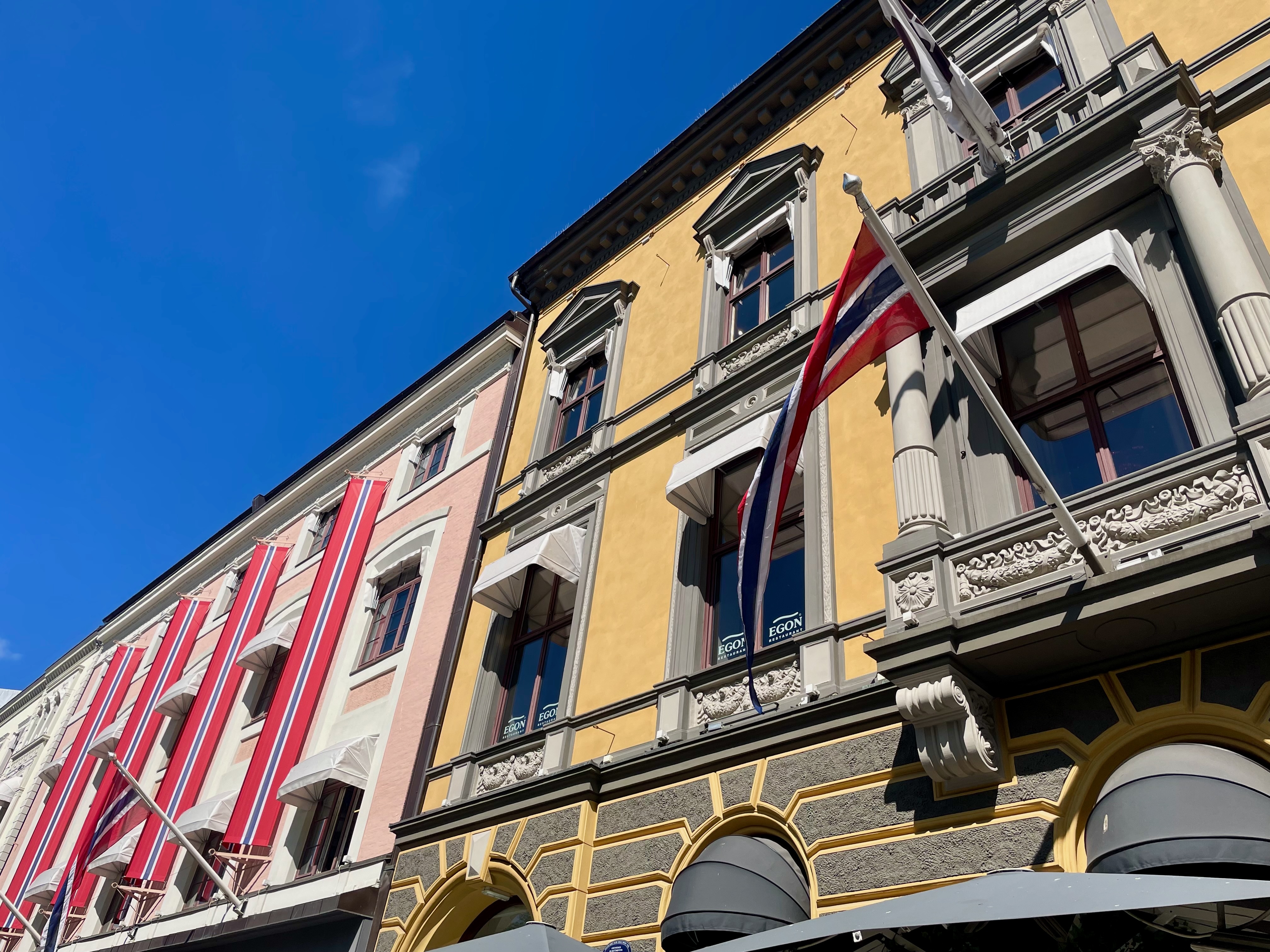 Egon Karl Johan historic yellow facade in Oslo with Norwegian flag, decorative windows, awnings, terrace umbrellas, and bright blue sky above Karl Johans gate.