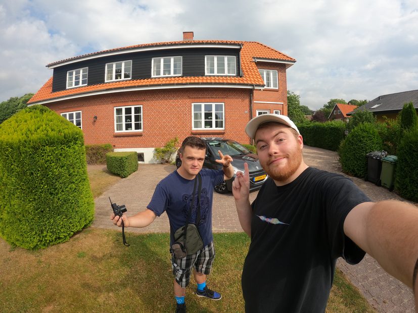 Kollund Rooms Review header photo showing two travelers in front of the red brick guesthouse in Kollund Østerskov, Denmark, with a Peugeot 208 parked on the driveway.