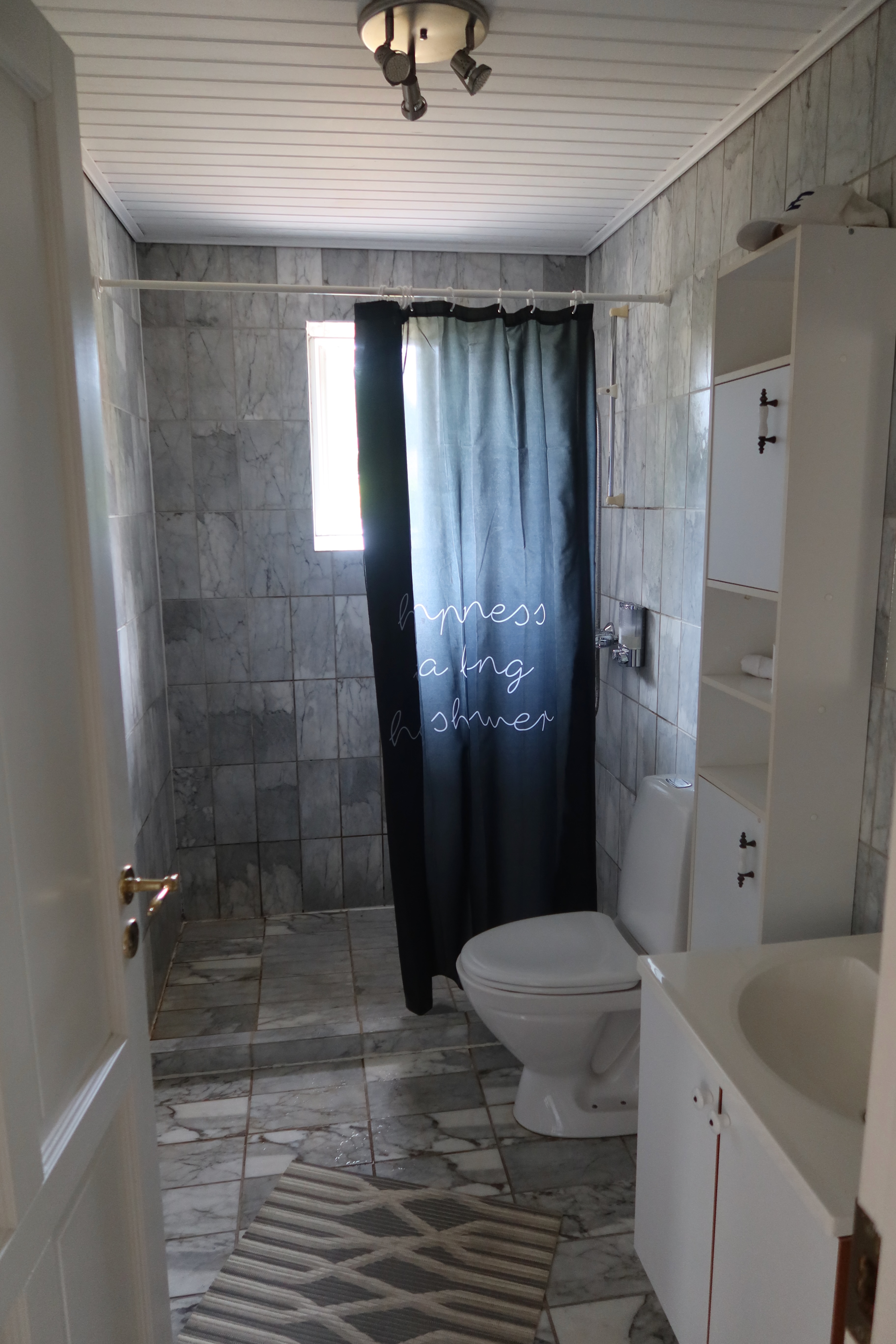 Kollund Rooms Review IMG 2597 showing the private bathroom with gray tiled shower area, black shower curtain, toilet, sink, storage cabinet, towels, and a simple guesthouse bathroom setup.