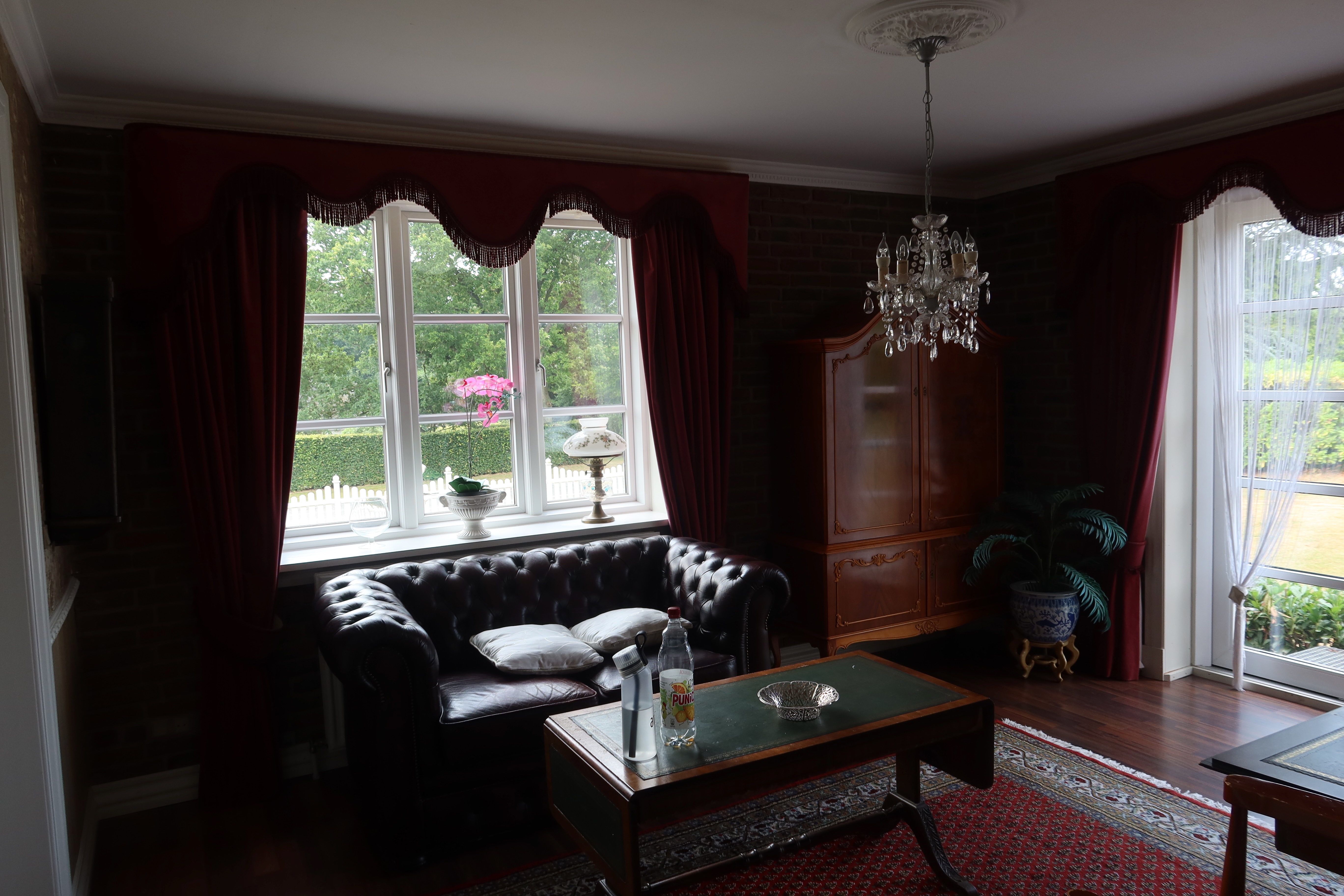 Kollund Rooms Review photo showing a cozy vintage sitting room with red curtains, a black leather sofa, chandelier, wooden cabinet, coffee table, and classic guesthouse decor in Denmark.