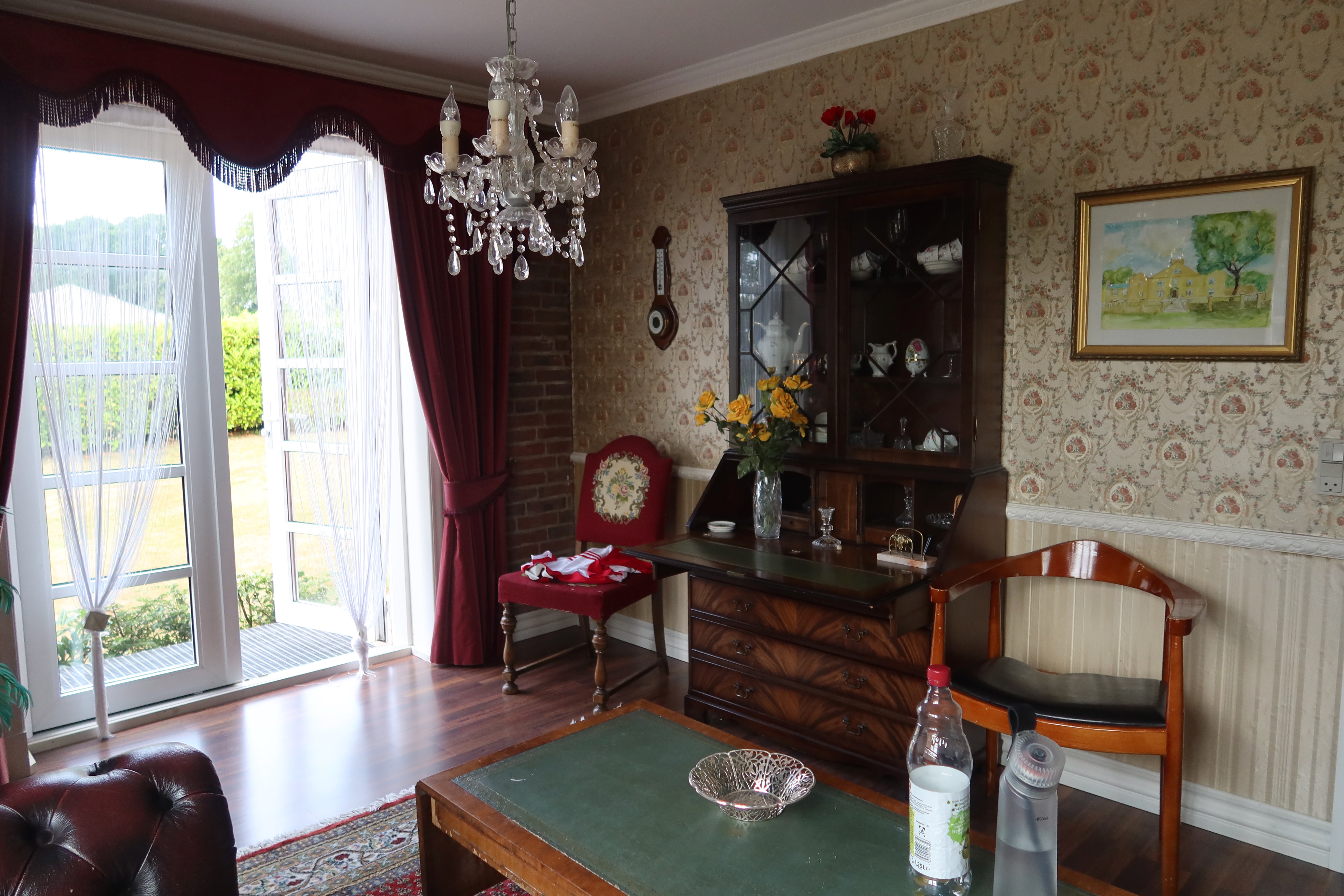 Kollund Rooms Review IMG 2599 showing a vintage shared lounge with red curtains, chandelier, floral wallpaper, wooden cabinet, leather sofa, and classic guesthouse decor in Denmark.