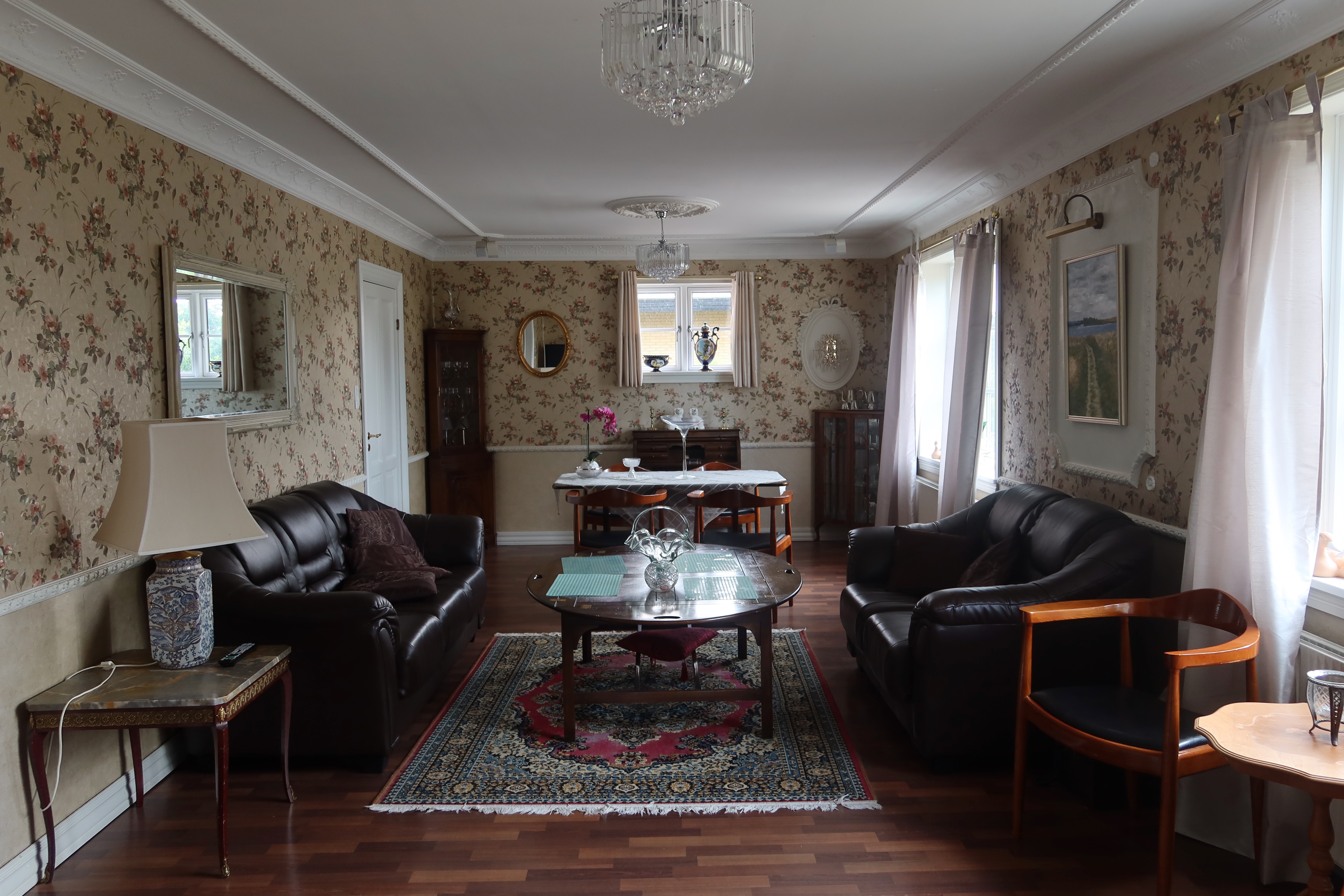 Kollund Rooms Review IMG 2600 showing the shared living room with black leather sofas, floral wallpaper, chandeliers, dining table, wooden furniture, and classic Danish guesthouse atmosphere.