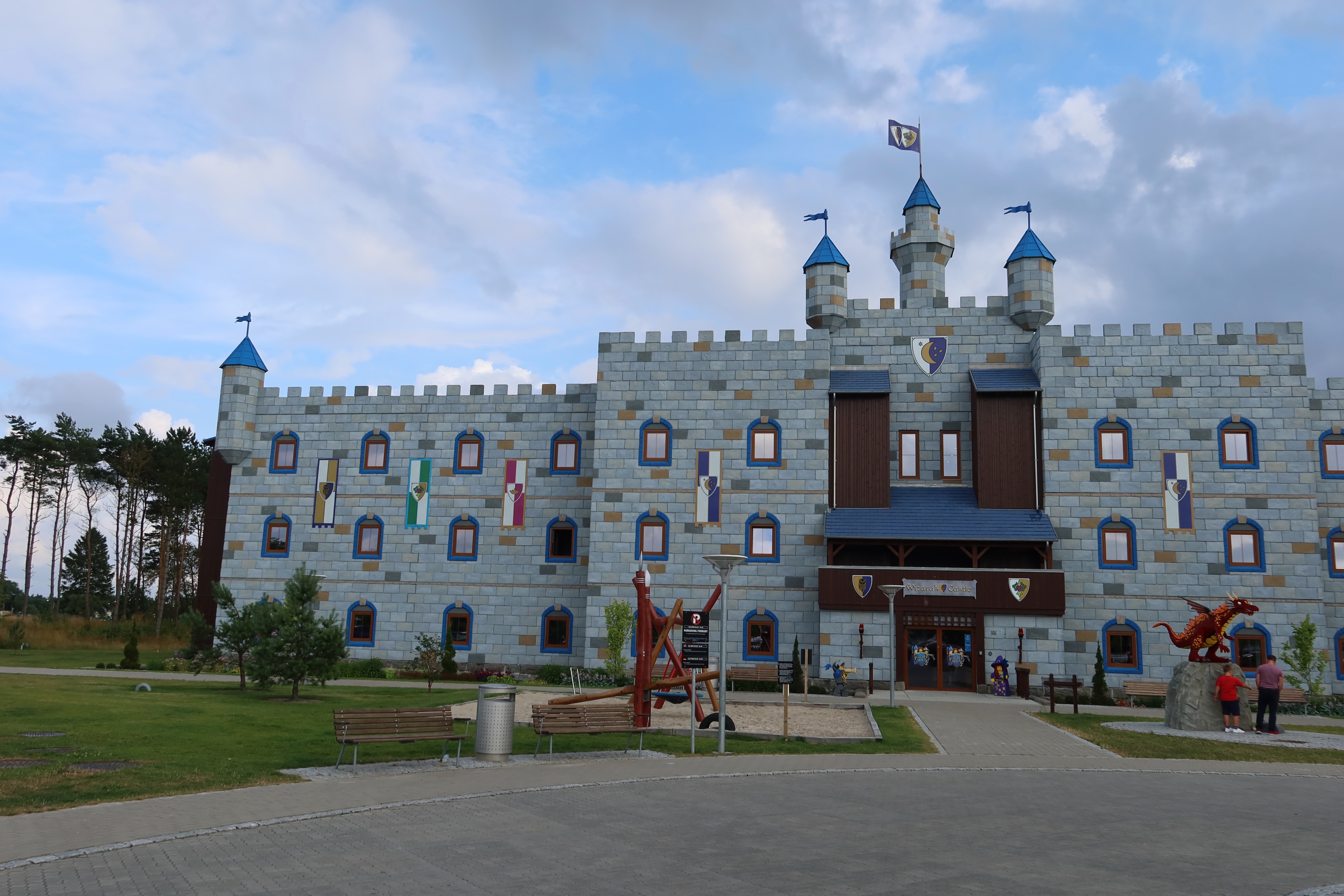 LEGOLAND Castle Hotel IMG 2878 showing the main Wizards Castle hotel building with castle towers blue rooftops flags playground benches and cloudy sky at LEGOLAND Billund Resort in Denmark