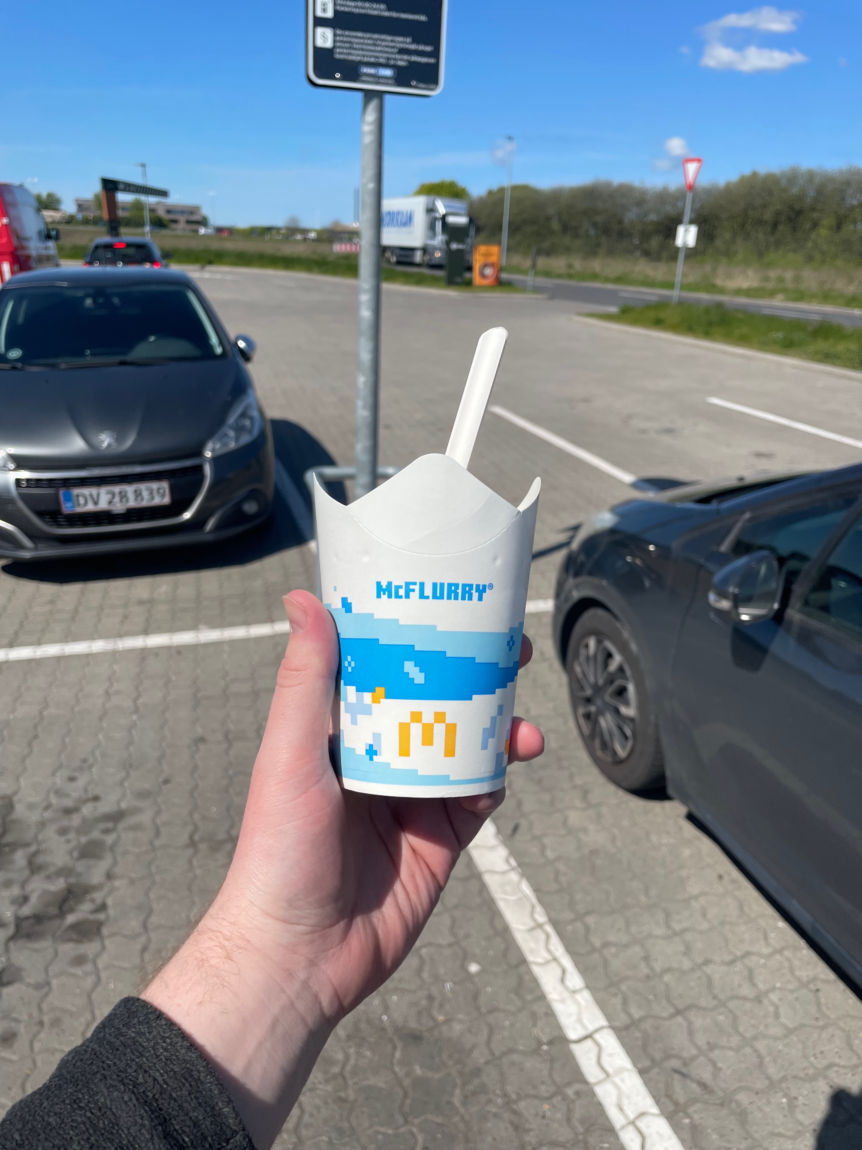 McDonald's Brønderslev McFlurry held outside in the parking lot on a sunny road trip stop in Denmark, with cars, blue sky and the restaurant area in the background
