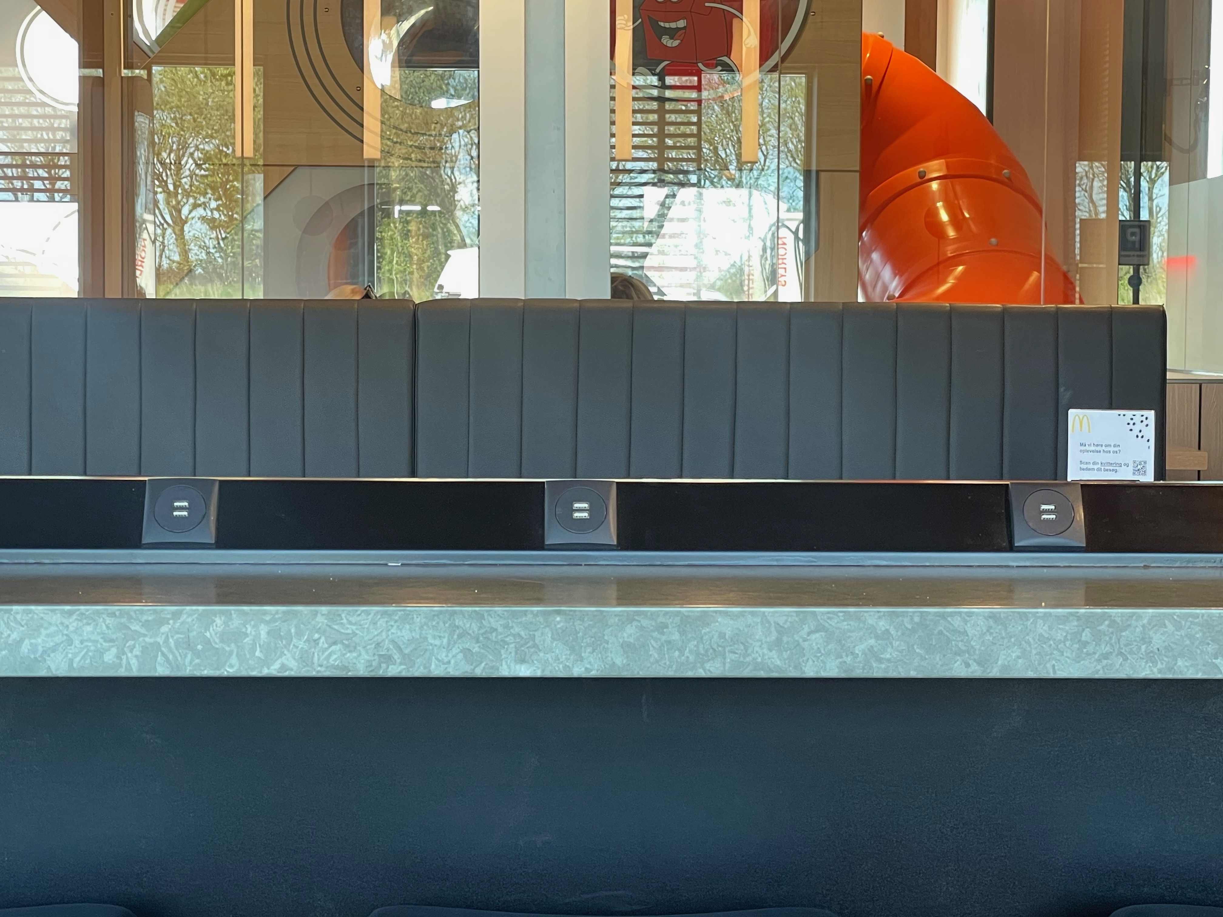 McDonald's Brønderslev seating area with built in USB charging points, black booth seating, glass play area view and orange indoor slide in Denmark