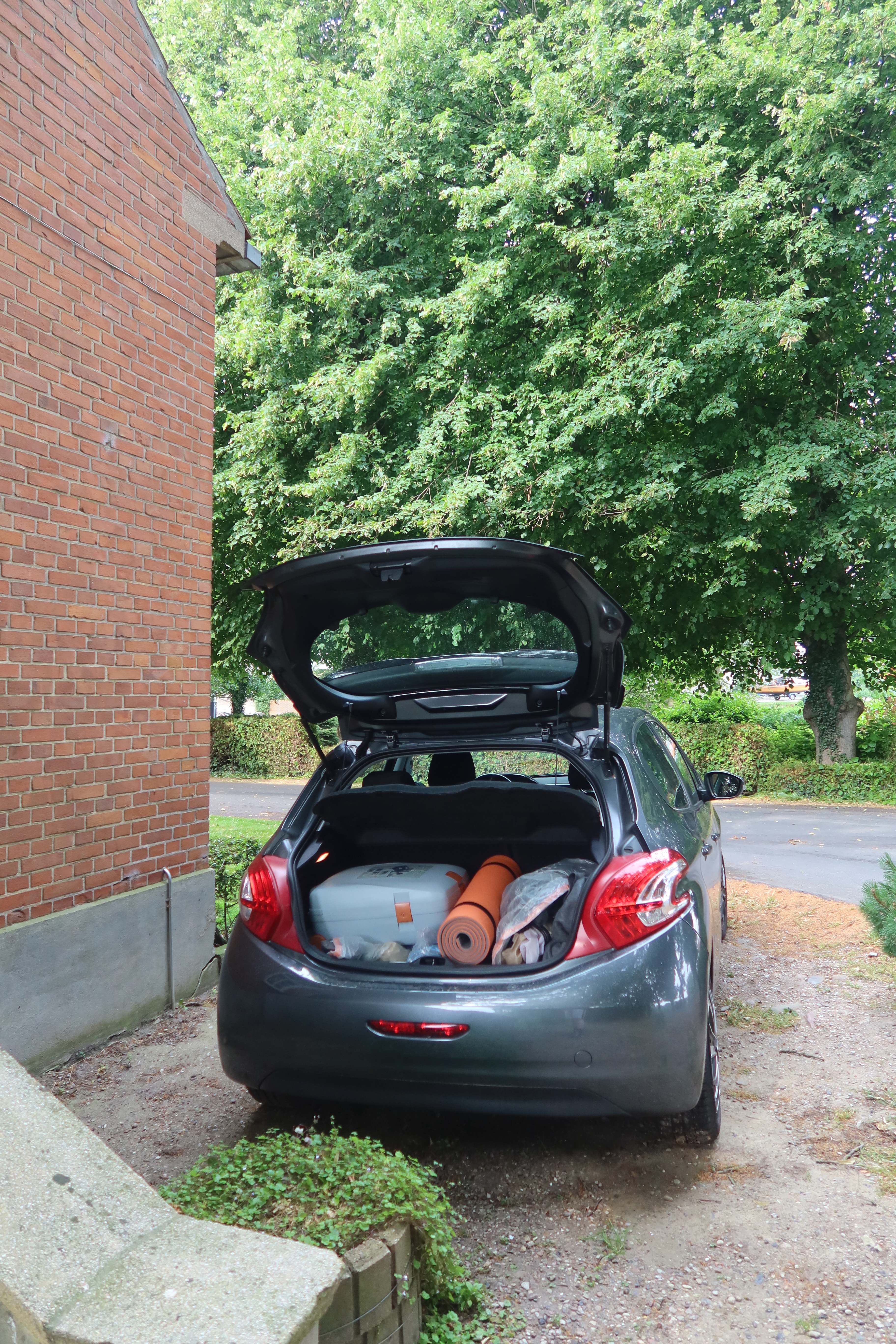 Mjelsgaard 1 Airbnb Review showing a gray Peugeot 208 parked beside the red brick Airbnb house in Denmark, with the trunk open and road trip camping gear inside