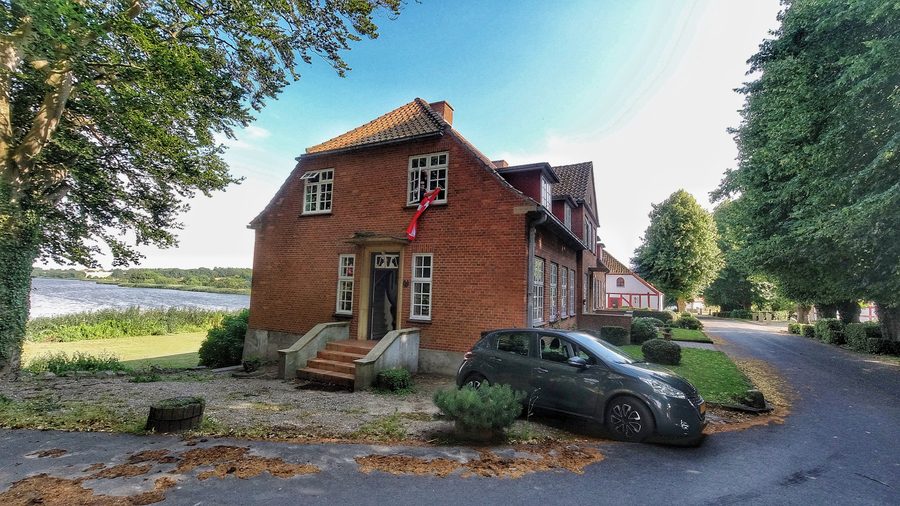 Mjelsgaard 1 Airbnb Review showing the red brick Airbnb house near Mjels Sø in Denmark with a gray Peugeot 208 parked outside and water views behind the property