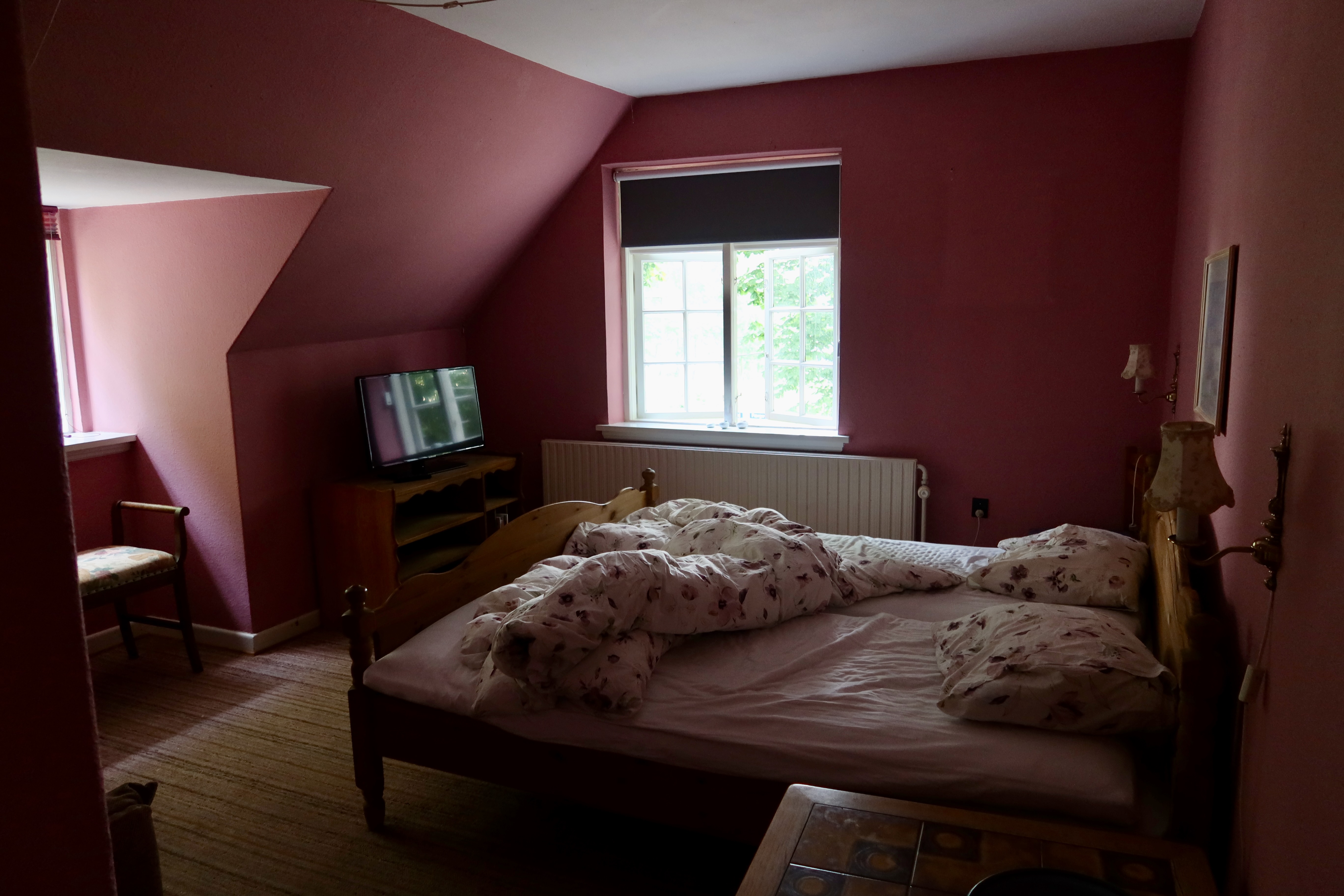 Mjelsgaard 1 Airbnb Review showing the simple pink Airbnb bedroom in Denmark with a wooden bed, floral bedding, a small television, and daylight through the window