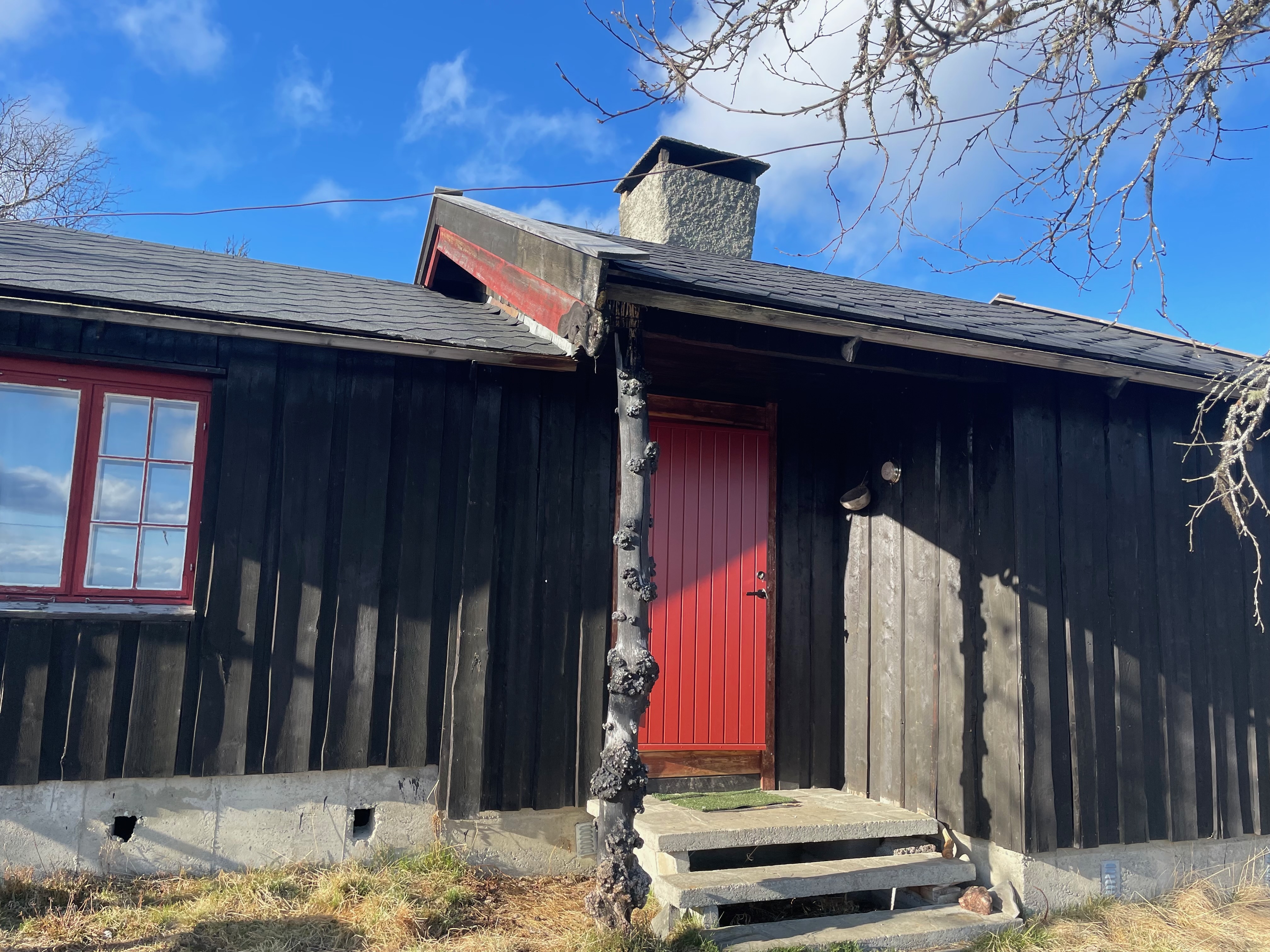 Per’s stuggu Sør-Fron black wooden cabin exterior with red door, red window frame, stone steps, chimney, dark timber walls and blue Norwegian sky.