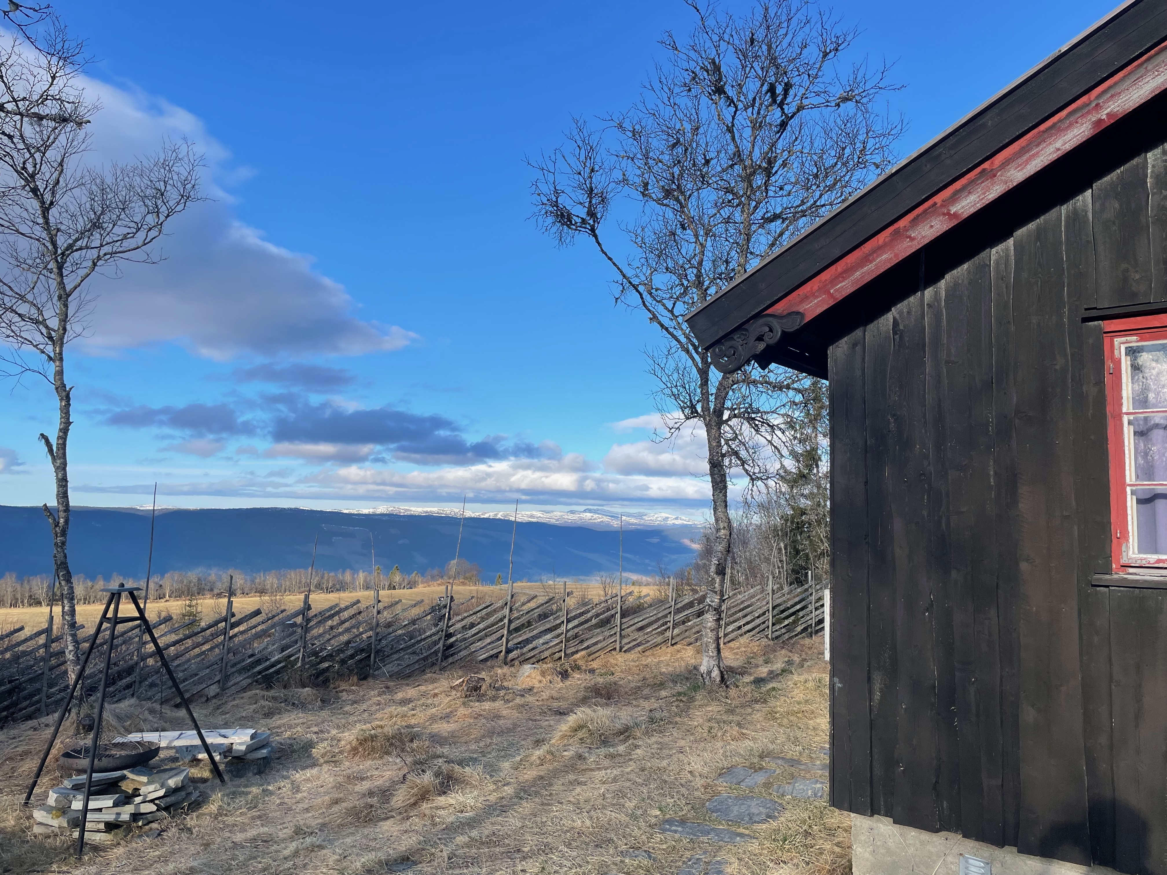 Per’s stuggu Sør-Fron cabin view with black wooden exterior, red window frame, outdoor fire pit, split rail fence, bare mountain trees and snowy Norwegian peaks in the distance.