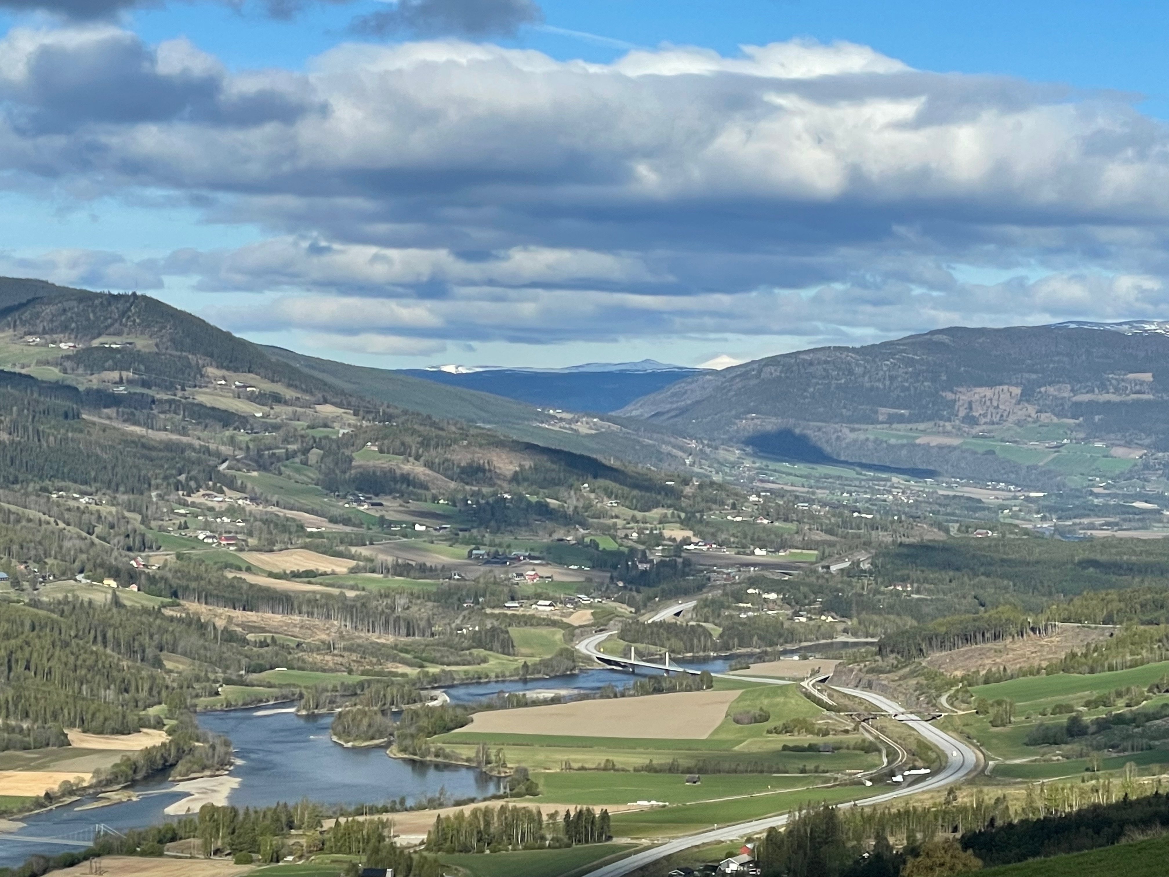 Per’s stuggu Sør-Fron mountain valley view while driving down from the cabin, with winding roads, river, farms, green fields, forested hills and cloudy Norwegian sky.