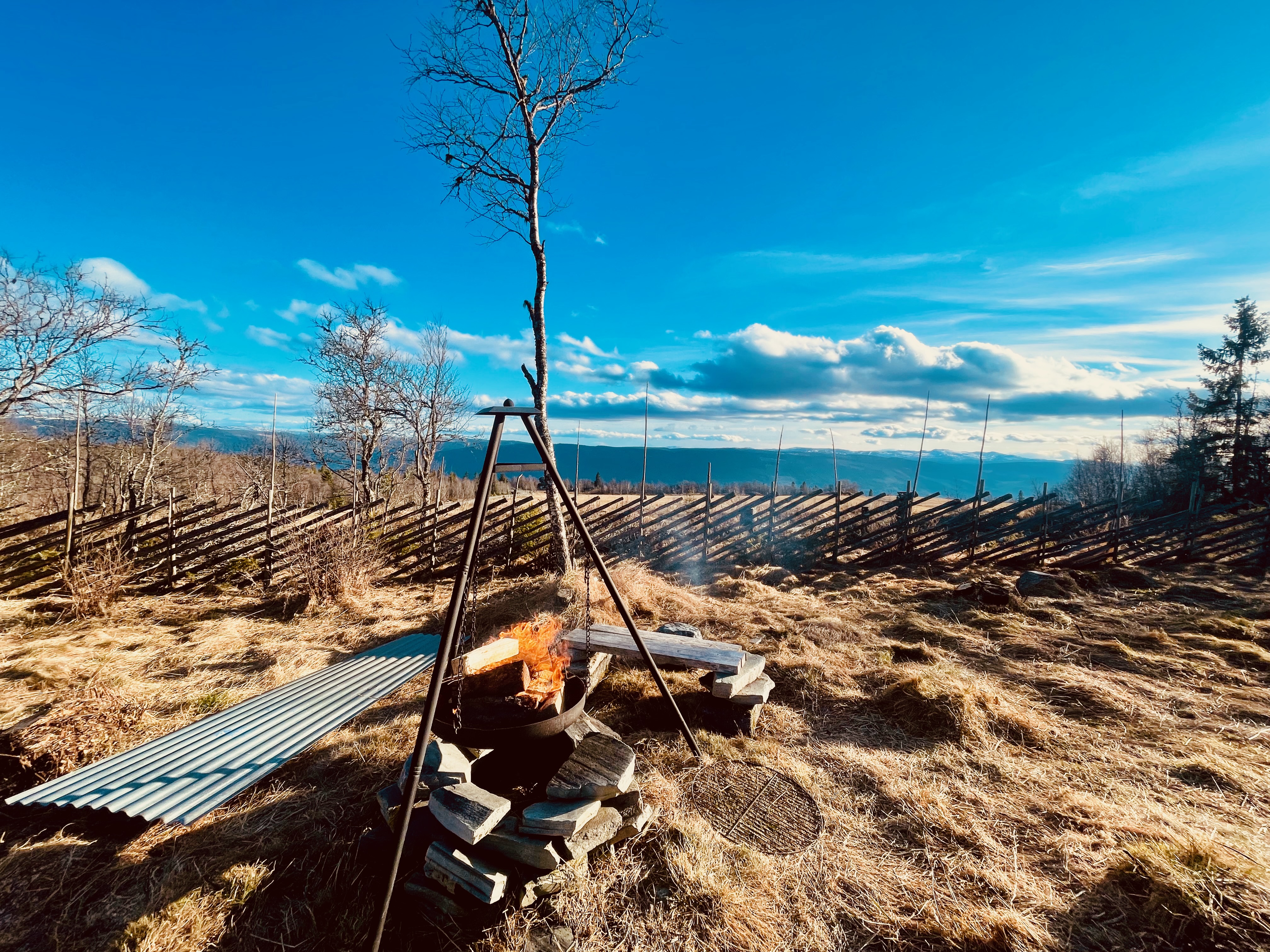 Per’s stuggu Sør-Fron outdoor fire pit with burning wood, black tripod grill, dry mountain grass, wooden fence, bare trees and wide blue sky over the Norwegian hills.