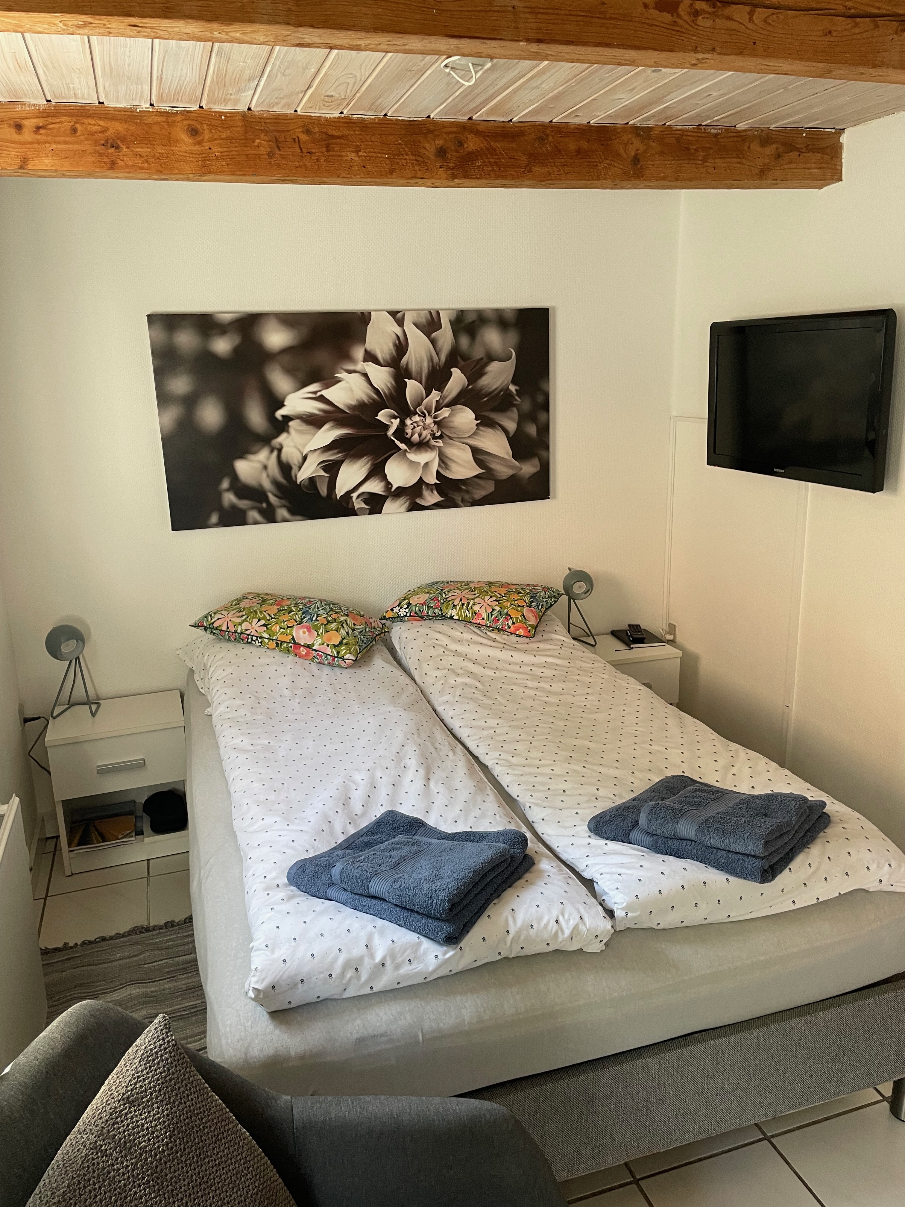 Private Annex Haderslev Review showing the cozy bedroom inside the Airbnb annex in Haderslev, with a double bed, folded towels, wall mounted TV, bedside tables, flower wall art and wooden ceiling beams.