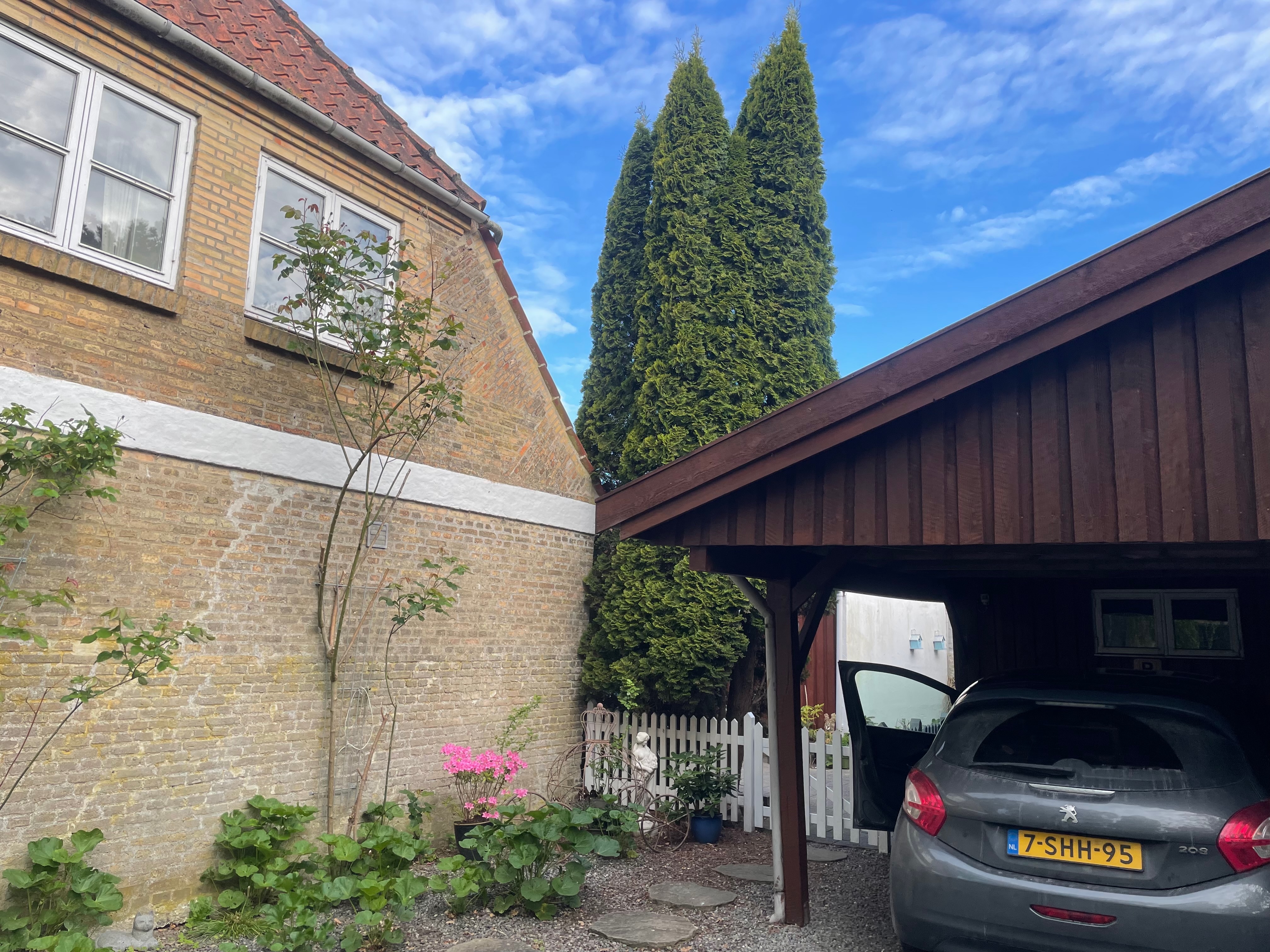 Private Annex Haderslev Review showing the private carport parking area in Haderslev with a parked Peugeot 208, brick house wall, garden plants, white fence and tall trees.
