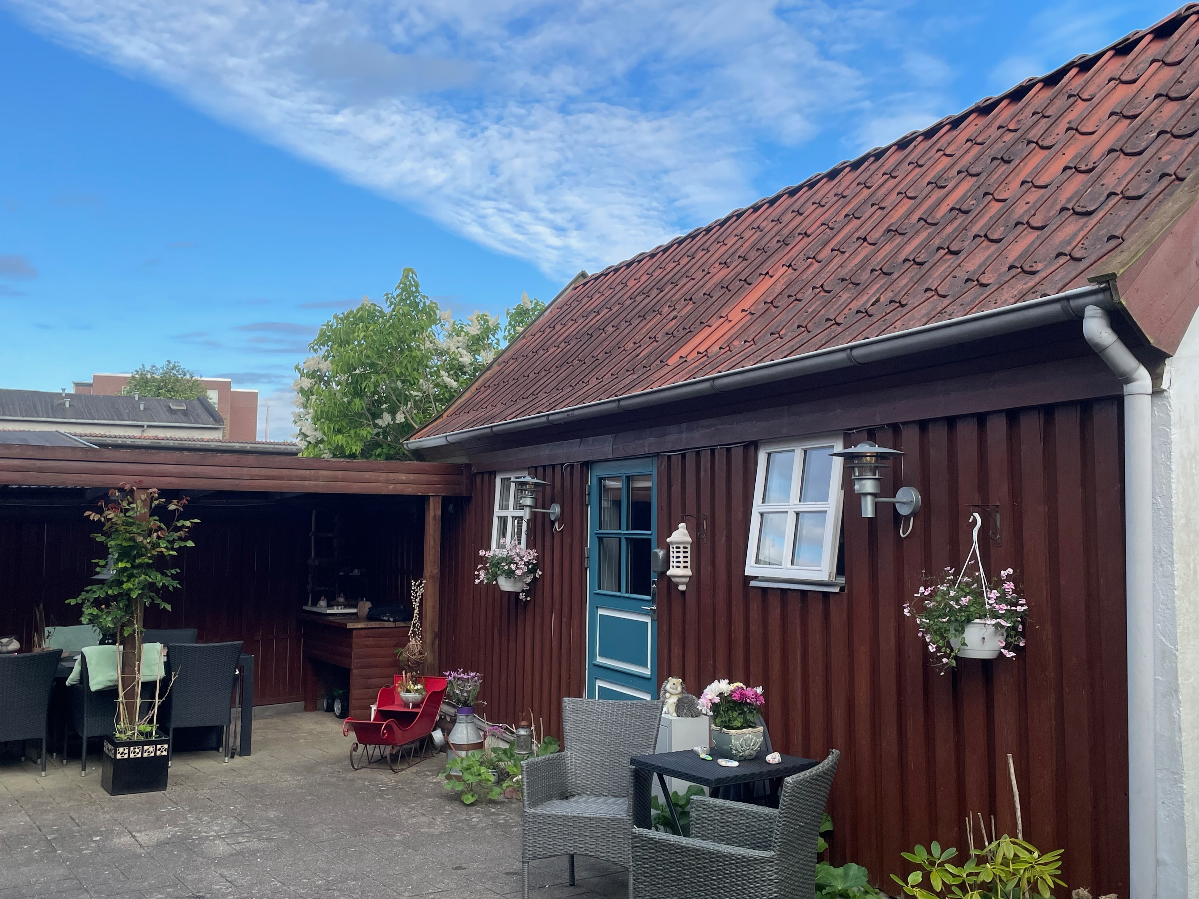 Private Annex Haderslev Review showing the charming Airbnb garden courtyard in Haderslev with a wooden annex, blue door, red tiled roof, outdoor seating, flowers and covered terrace.