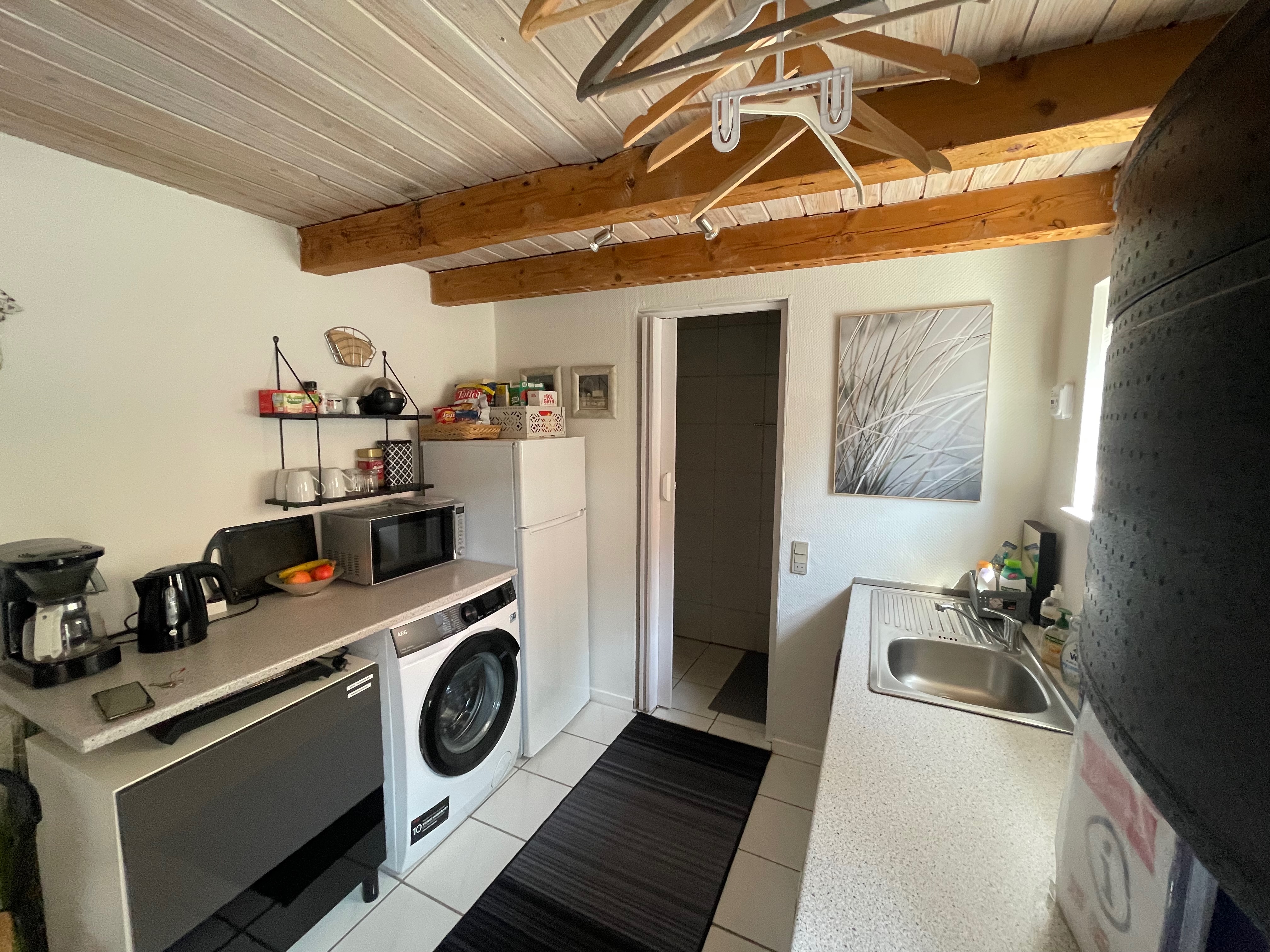 Private Annex Haderslev Review showing the compact mini kitchen inside the Airbnb annex in Haderslev, with AEG washing machine, microwave, fridge, freezer, coffee maker, kettle, sink, snacks and wooden ceiling beams.