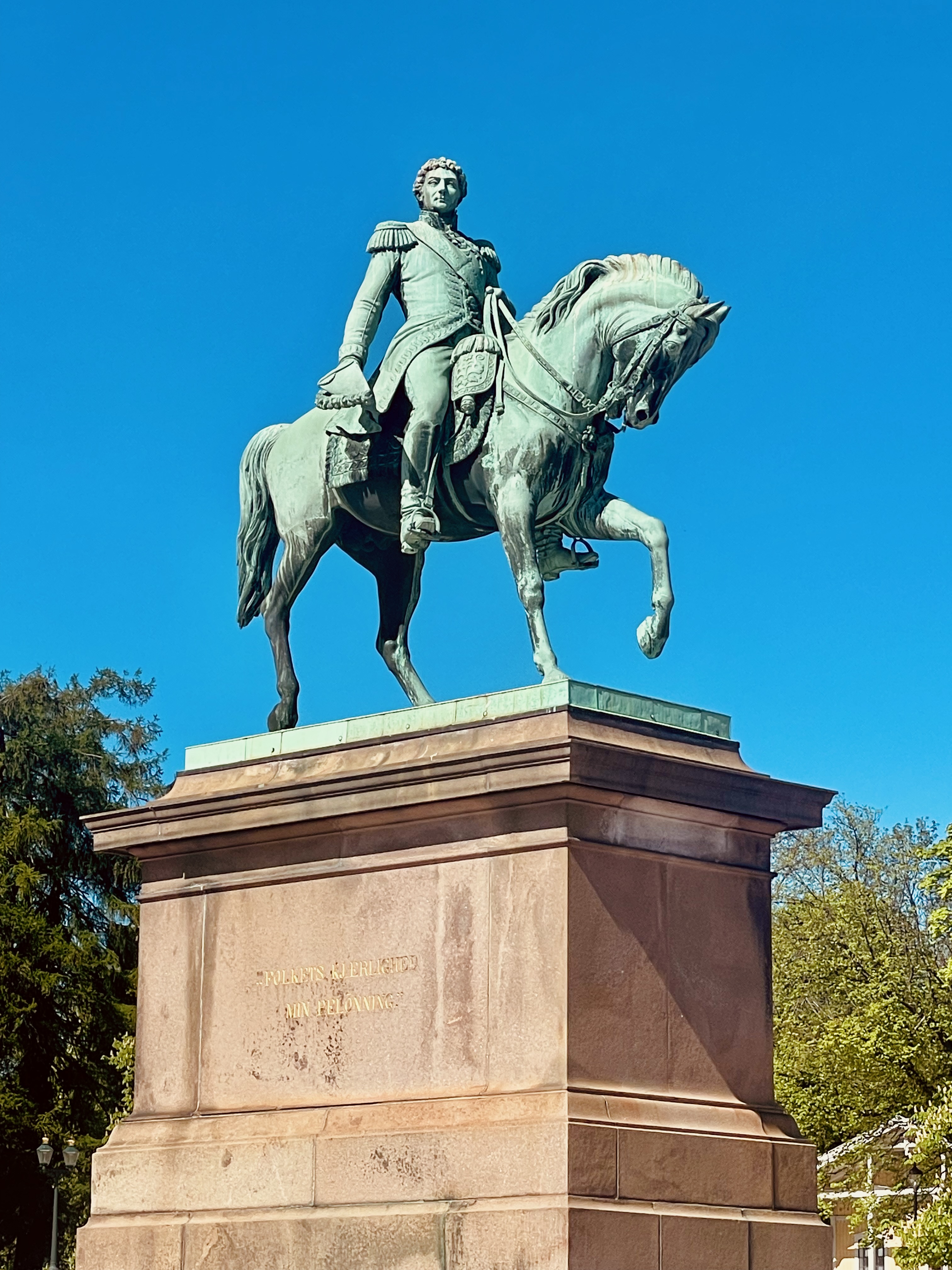 Royal Palace Oslo detail with the equestrian statue of King Karl Johan on Slottsplassen, showing the bronze rider and horse statue against a clear blue sky.