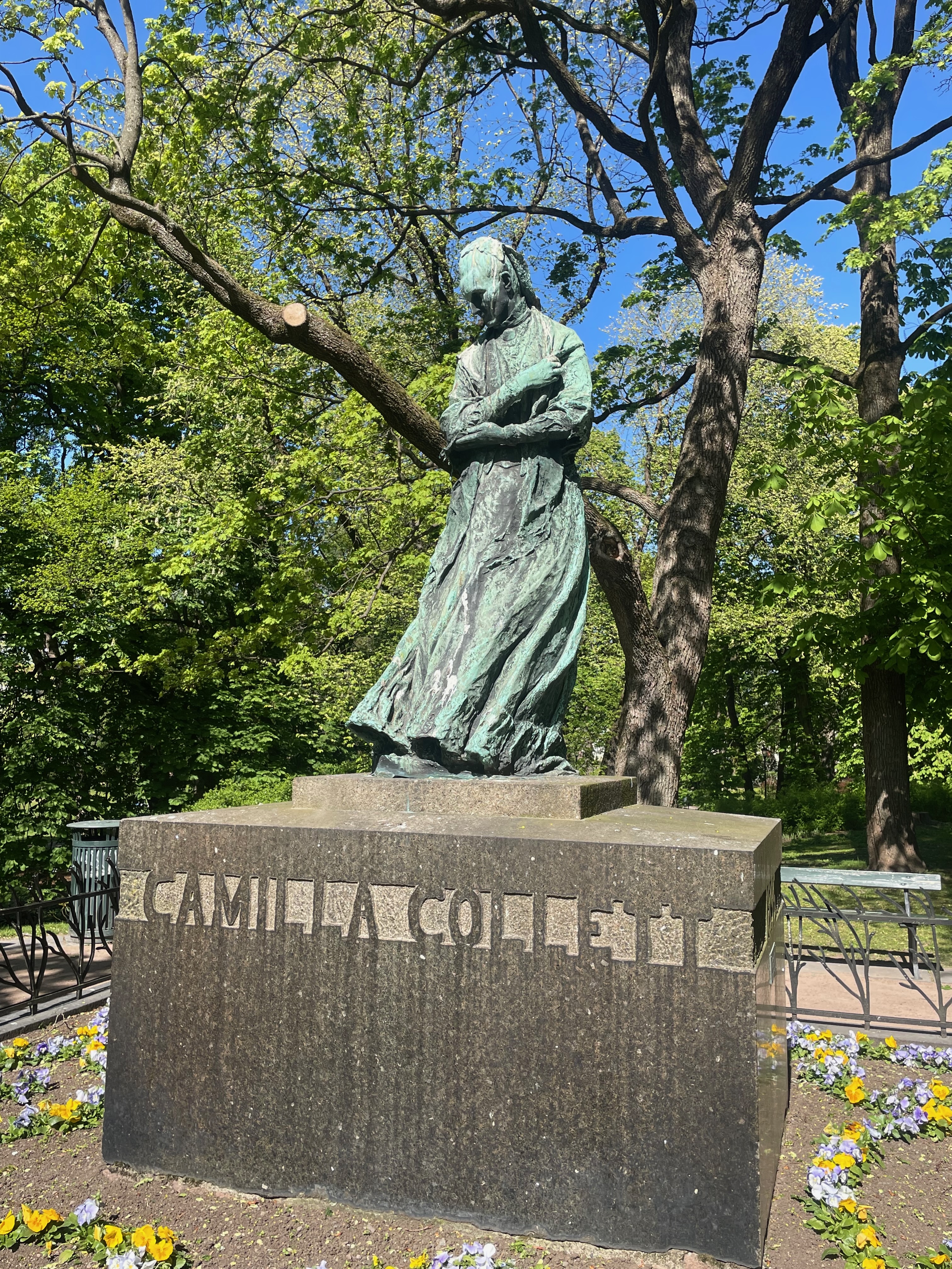 Royal Palace Oslo area in Slottsparken with the Camilla Collett statue surrounded by trees, flowers, benches, and green park scenery.