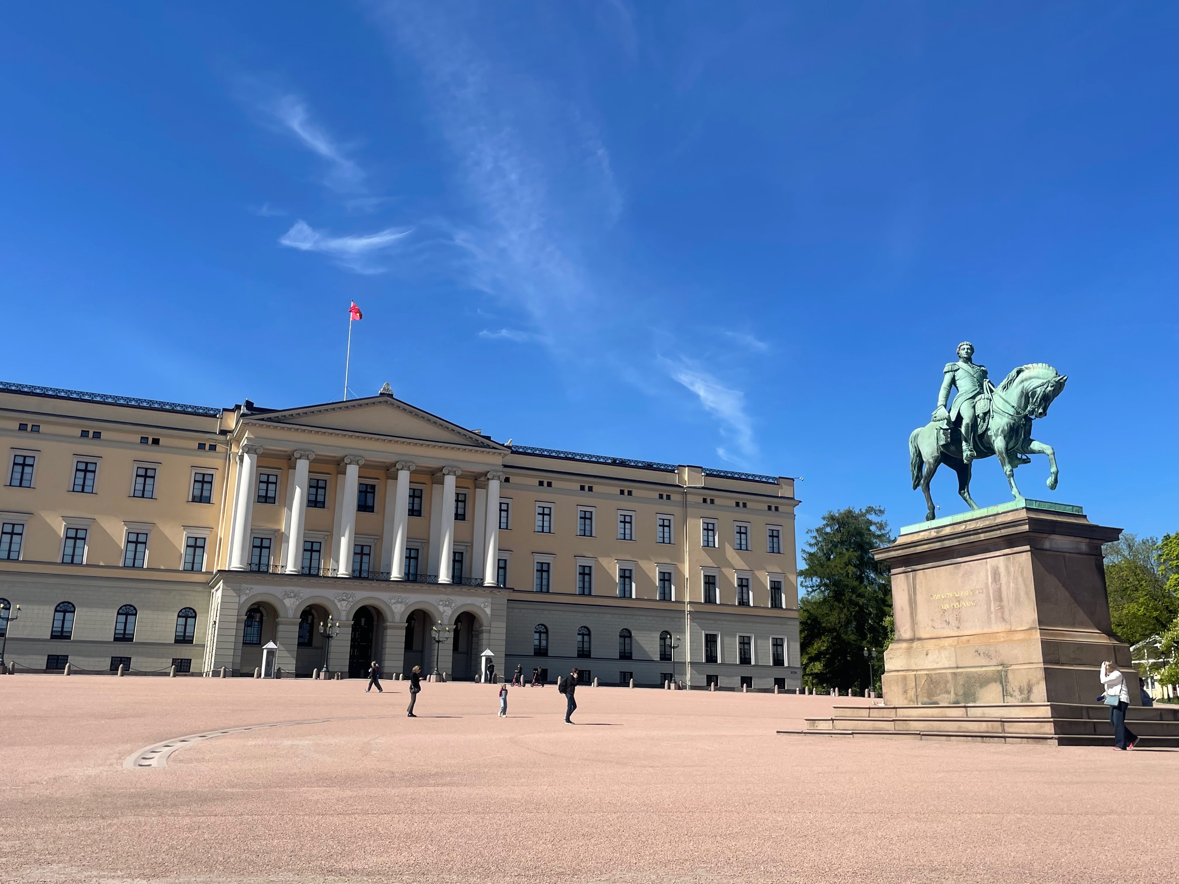 Royal Palace Oslo seen from Slottsplassen with Det kongelige slott, the wide palace square, visitors, the Norwegian flag, and the equestrian statue of King Karl Johan under a clear blue sky.