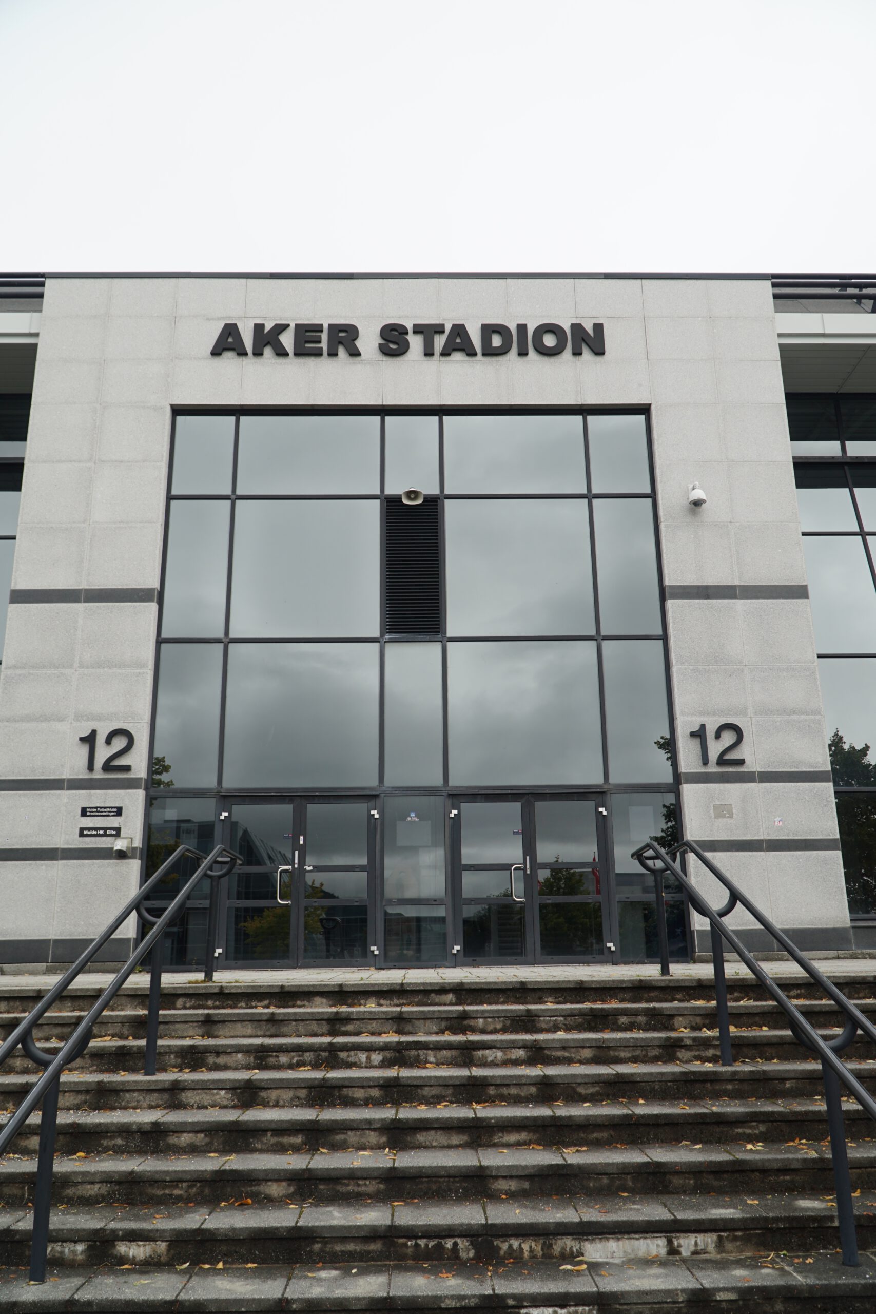 Aker Stadion Molde main entrance with large glass facade, staircase leading up to doors marked section 12, and the Aker Stadion sign above the entrance.