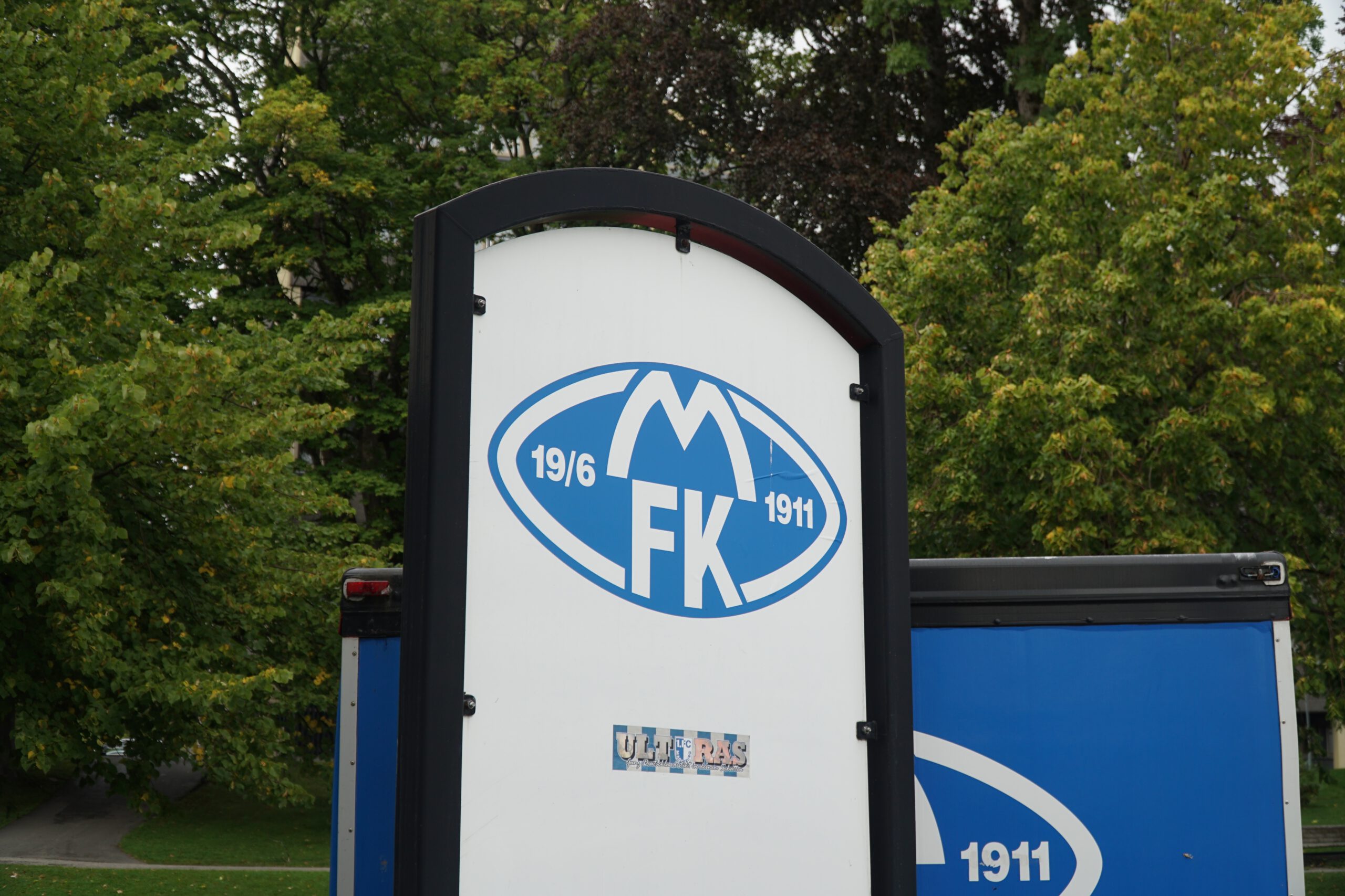 Aker Stadion Molde entrance sign featuring the Molde FK logo with founding date 1911, surrounded by green trees near the stadium grounds in Norway.