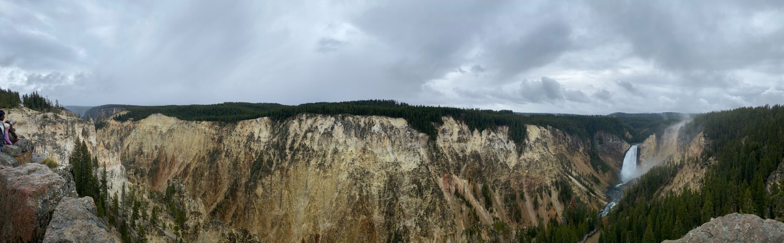Wide panoramic view of the Grand Canyon of the Yellowstone with the Lower Falls cascading into the canyon, showcasing dramatic cliffs, stormy skies, and the vast scale of Yellowstone National Park.