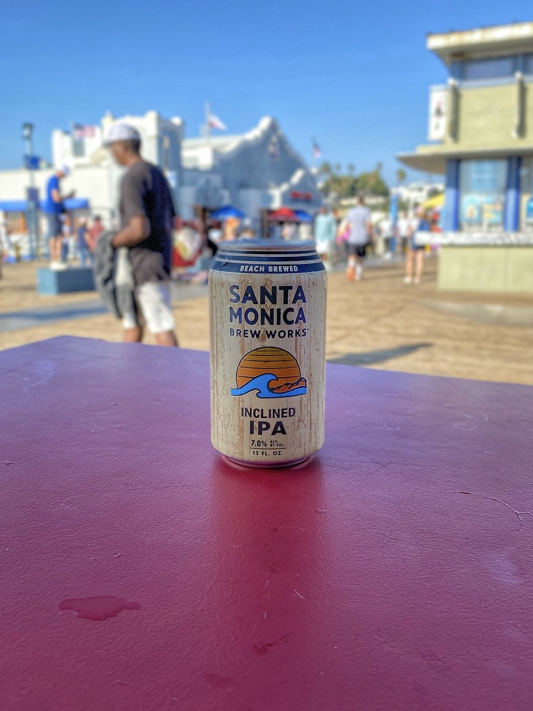 Santa Monica Brew Works Inclined IPA beer can on a red table at Santa Monica Pier, marking a first beer in America with beach crowds and Pacific Ocean atmosphere in the background on the PierBurger Santa Monica Pier area