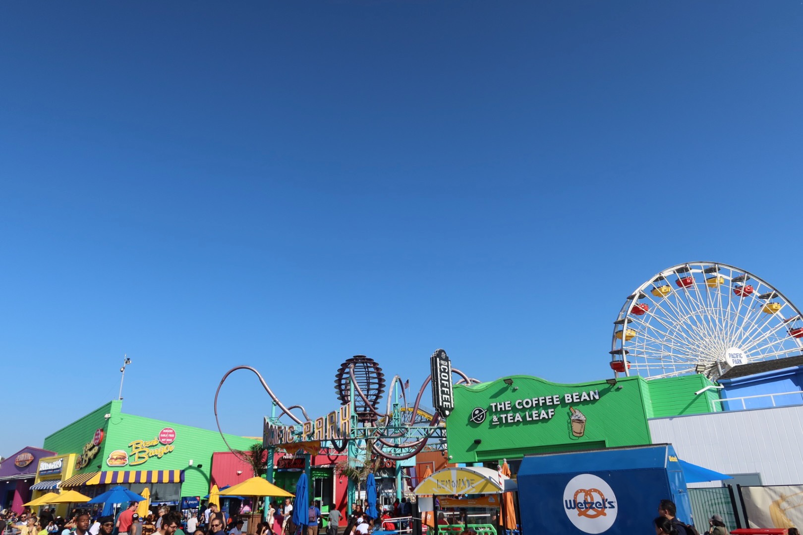 Busy Santa Monica Pier scene with colorful food stands, arcade signage, The Coffee Bean & Tea Leaf storefront, looping roller coaster tracks, and the Pacific Park Ferris wheel rising above the pier under a clear blue sky.