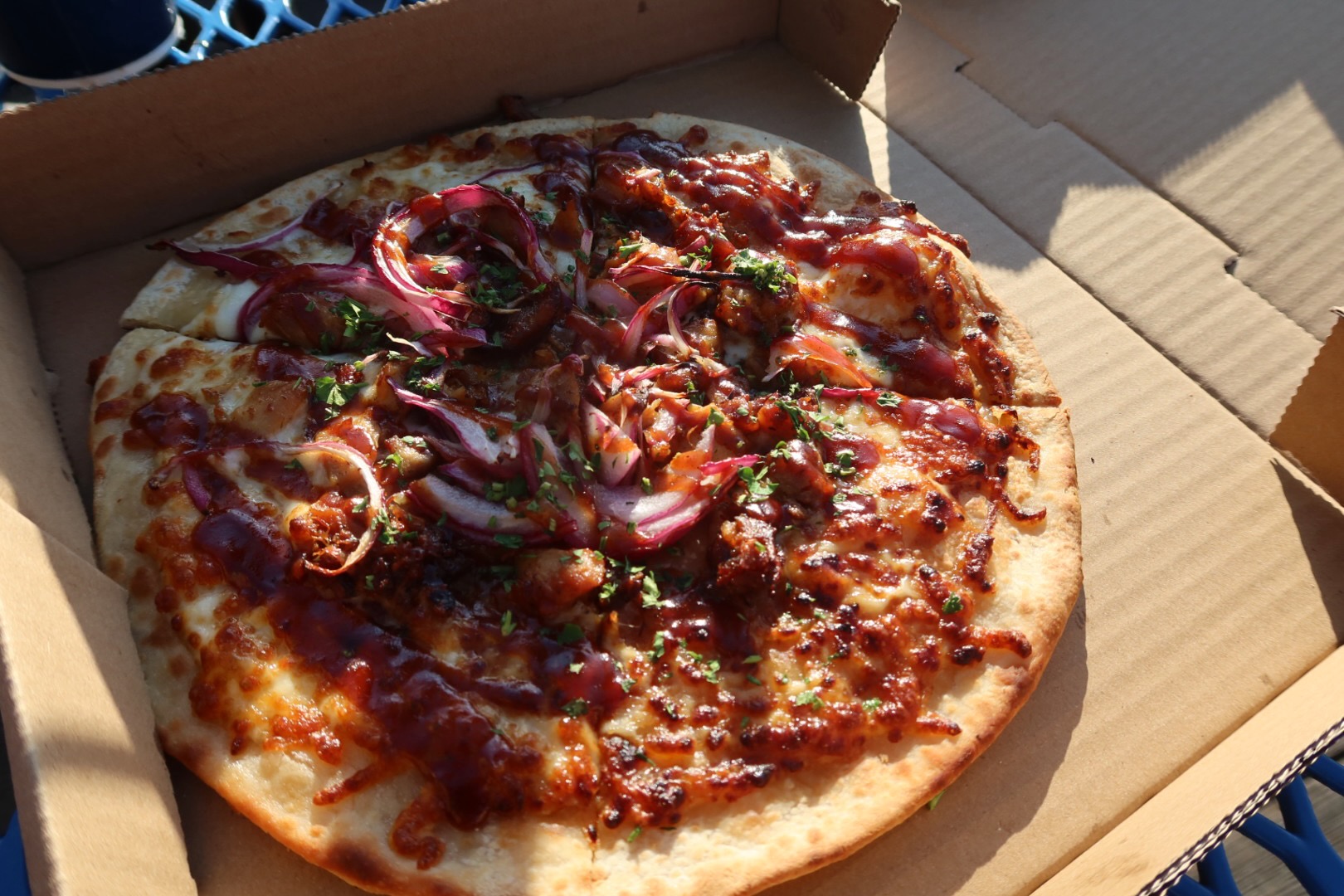 Barbecue chicken pizza from Mr. Nice Fry at Santa Monica Pier, topped with melted cheese, barbecue sauce, red onions and herbs, served fresh in an open cardboard pizza box at an outdoor table.