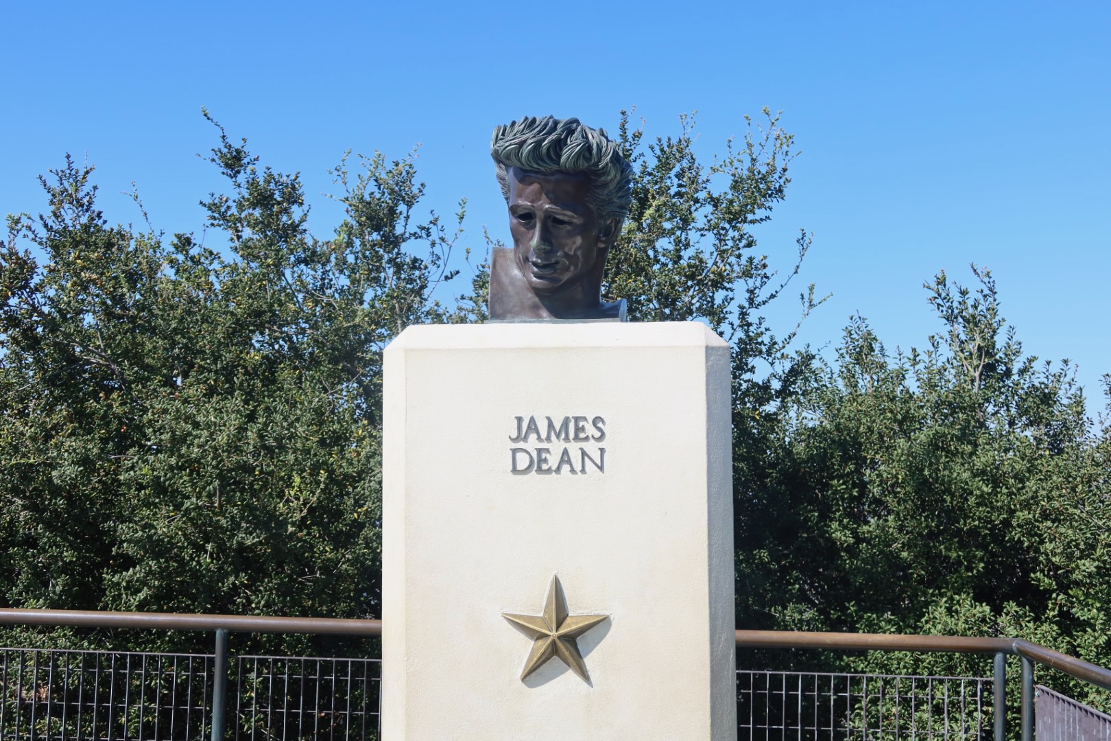 Bronze bust of James Dean on a white pedestal at Griffith Observatory, featuring a gold star emblem, surrounded by greenery under a clear blue sky in Griffith Park, Los Angeles.