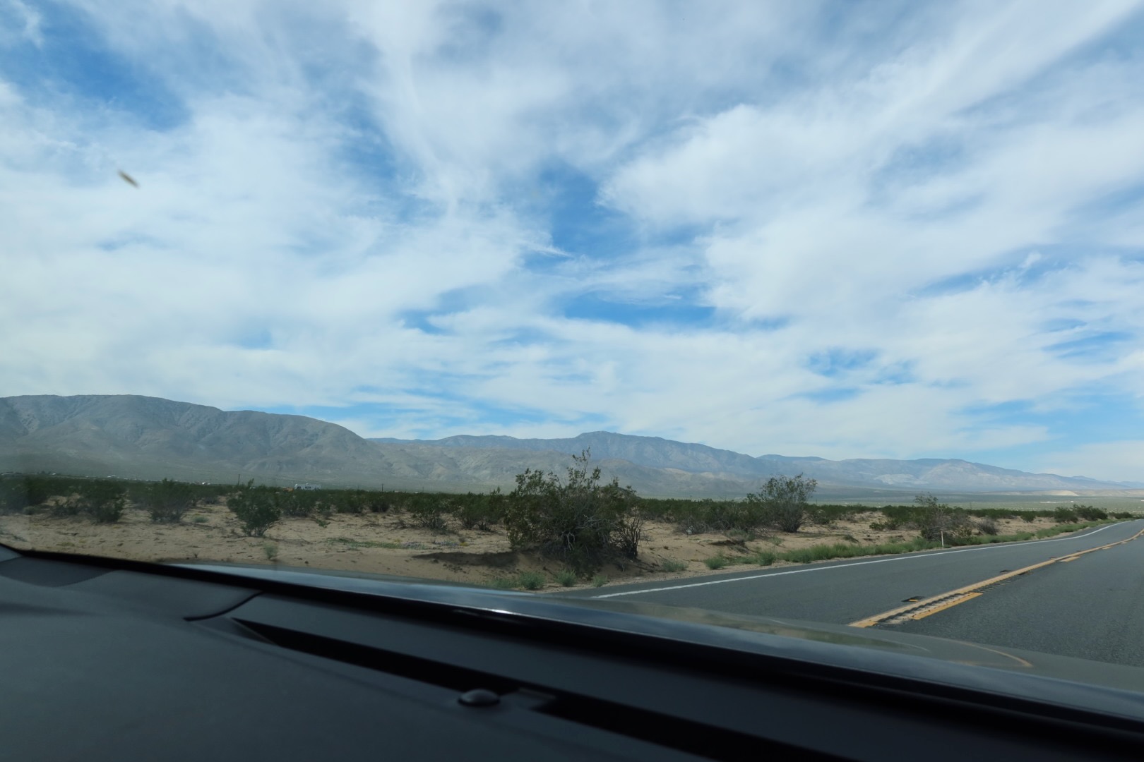 Driving through the Mojave Desert in California, seen from inside a car with a desert highway, low shrubs, and distant Mojave Desert mountains under a wide, cloud-filled sky.
