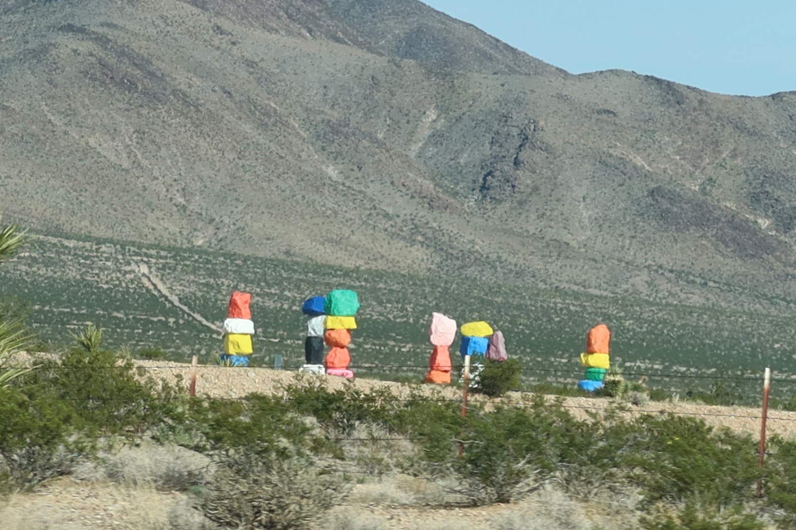 Seven Magic Mountains art installation with brightly colored stacked rock towers standing in the Mojave Desert near Interstate 15, photographed from a moving car with desert shrubs and mountain slopes in the background.