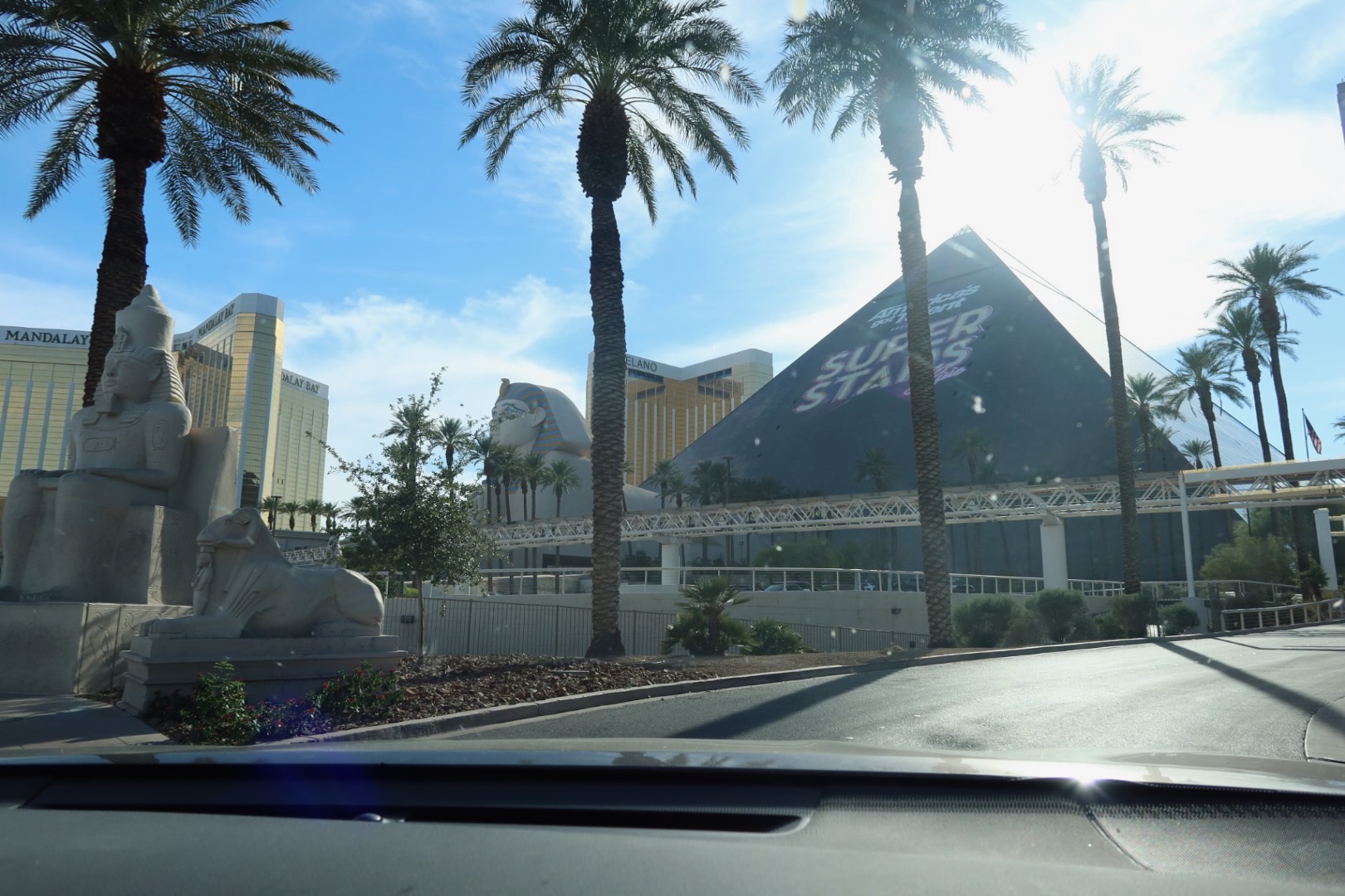 Daytime exterior view of the Luxor Hotel & Casino Las Vegas pyramid with palm trees, the Sphinx statue at the entrance, Mandalay Bay towers in the background, and the resort seen from the road in bright desert sunlight.