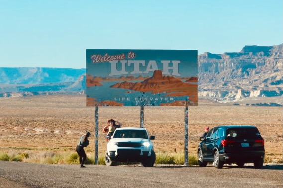 Welcome to Utah state sign near Glen Canyon, road trip stop on the Arizona Utah border with desert landscape, parked cars, and wide open Southwest scenery